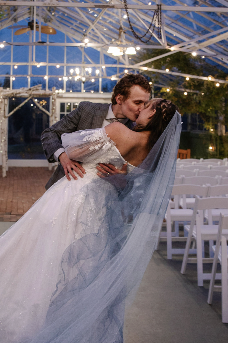 A bride and groom sharing a kiss at their wedding reception in a glass-roofed venue decorated with string lights.
