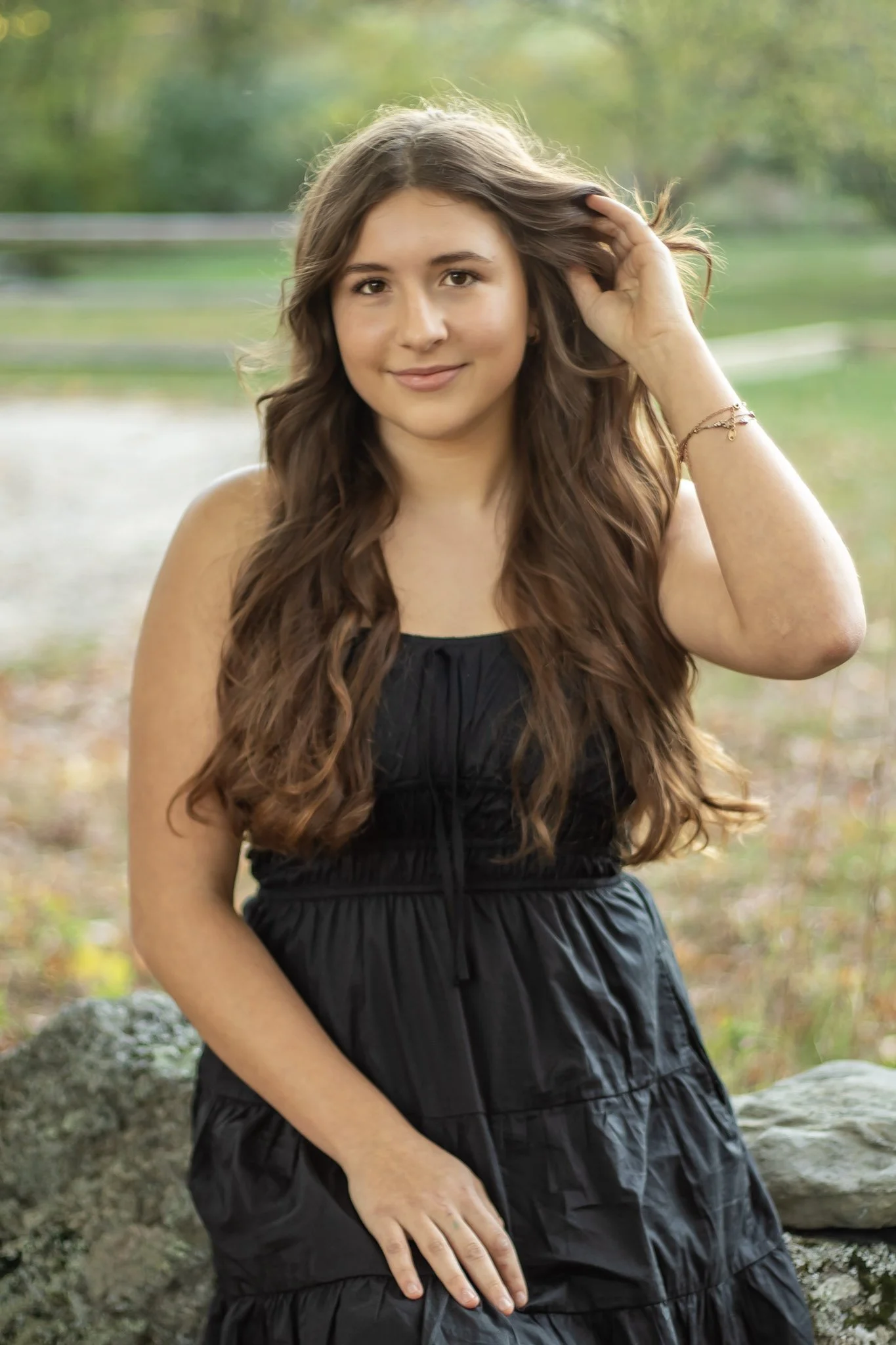 A young woman with long, wavy brown hair wearing a black sleeveless dress outdoors in a park.