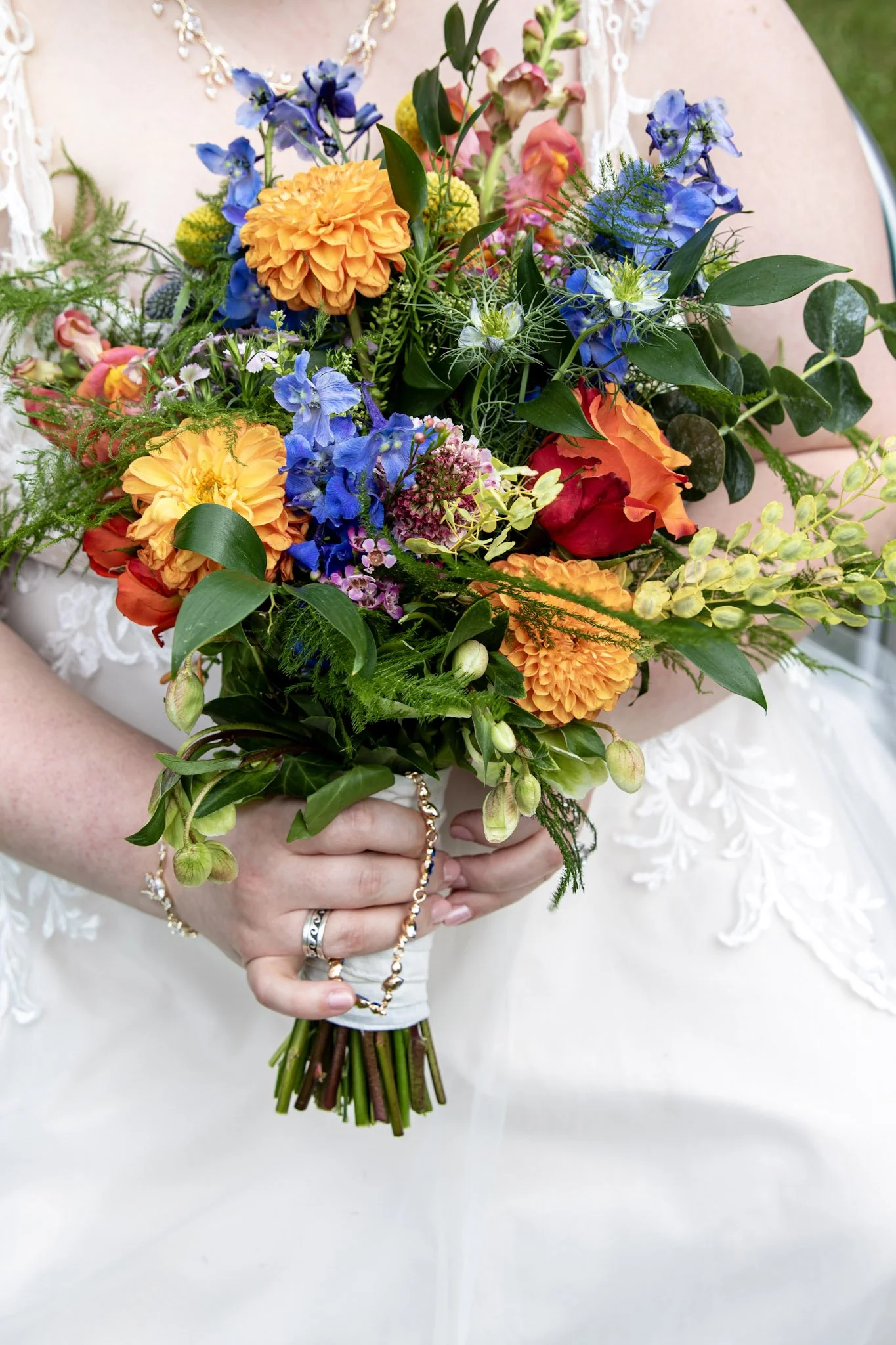 A person in a white dress holding a colorful bouquet of flowers, including orange, yellow, blue, and pink blooms, with their hands and jewelry visible.