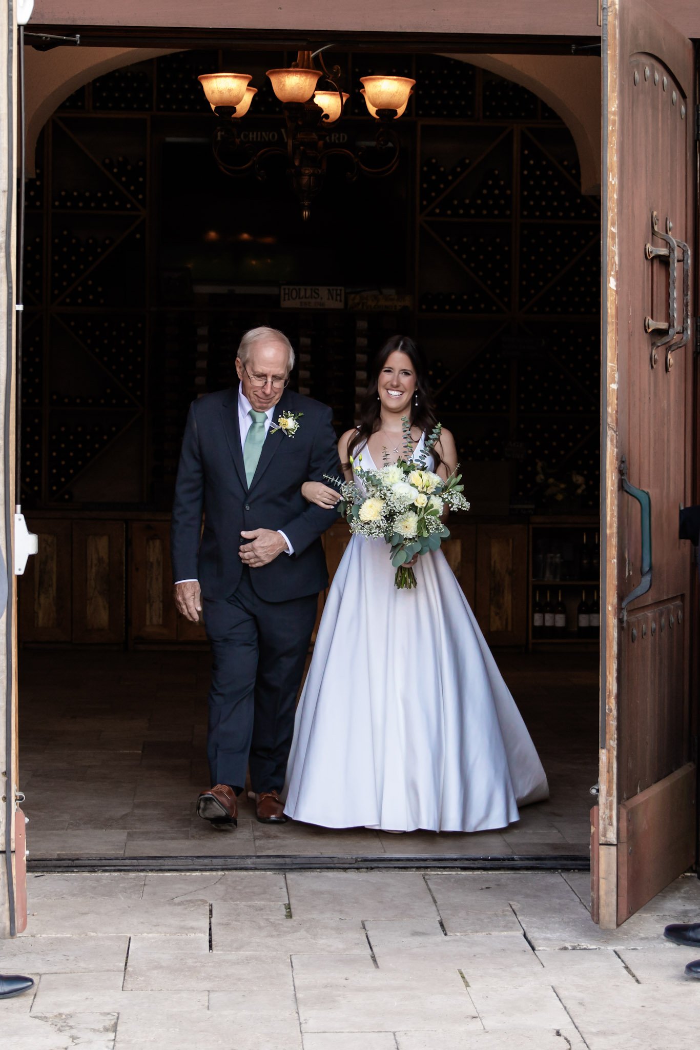 A bride walking down the aisle with an older man, likely her father, at a wedding. She is wearing a white wedding dress and holding a bouquet of white and yellow flowers. They are inside a rustic building with wooden doors open.