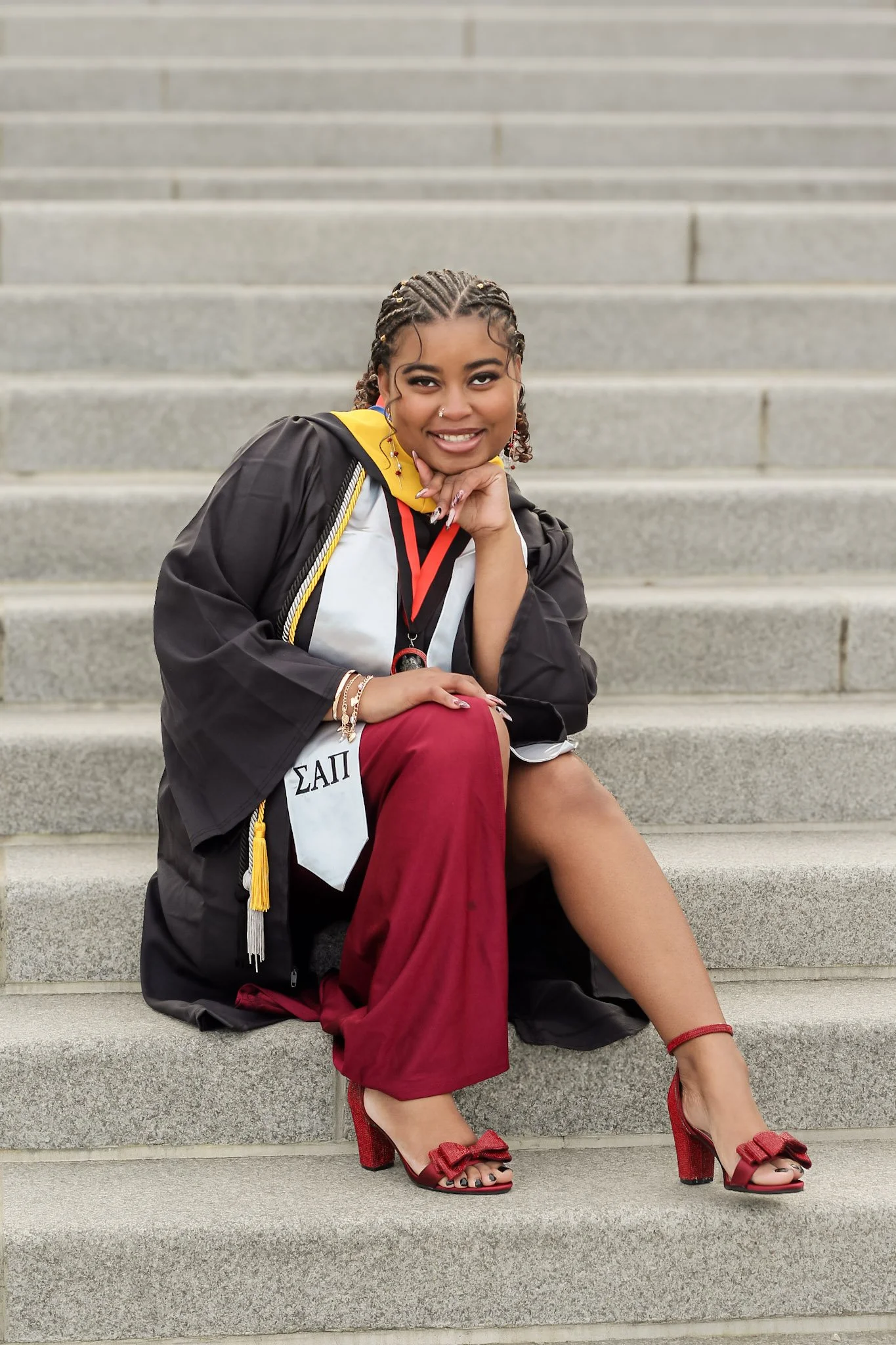 A young woman in graduation attire sitting on steps, wearing a black cap and gown with Greek letters, red pants, red heels, and jewelry.