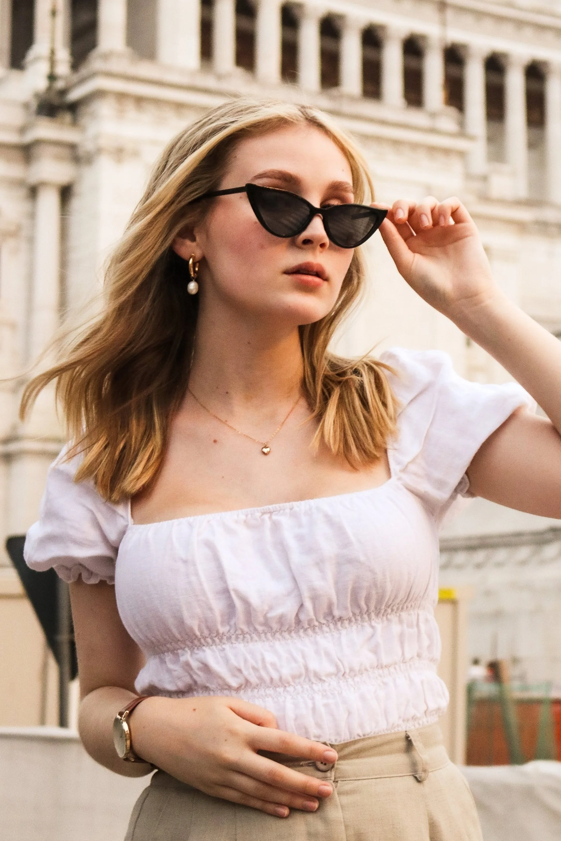 A young woman with blonde hair wearing black sunglasses, a white puff-sleeve top, beige pants, and jewelry, standing outdoors with architecture in the background.