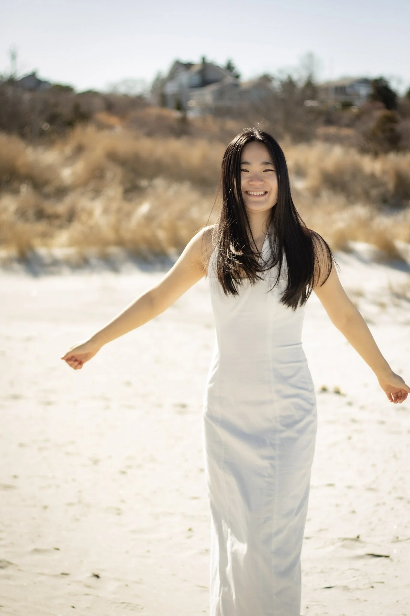 A young woman with long dark hair smiling and standing on a sandy beach with dry grass and houses in the background.