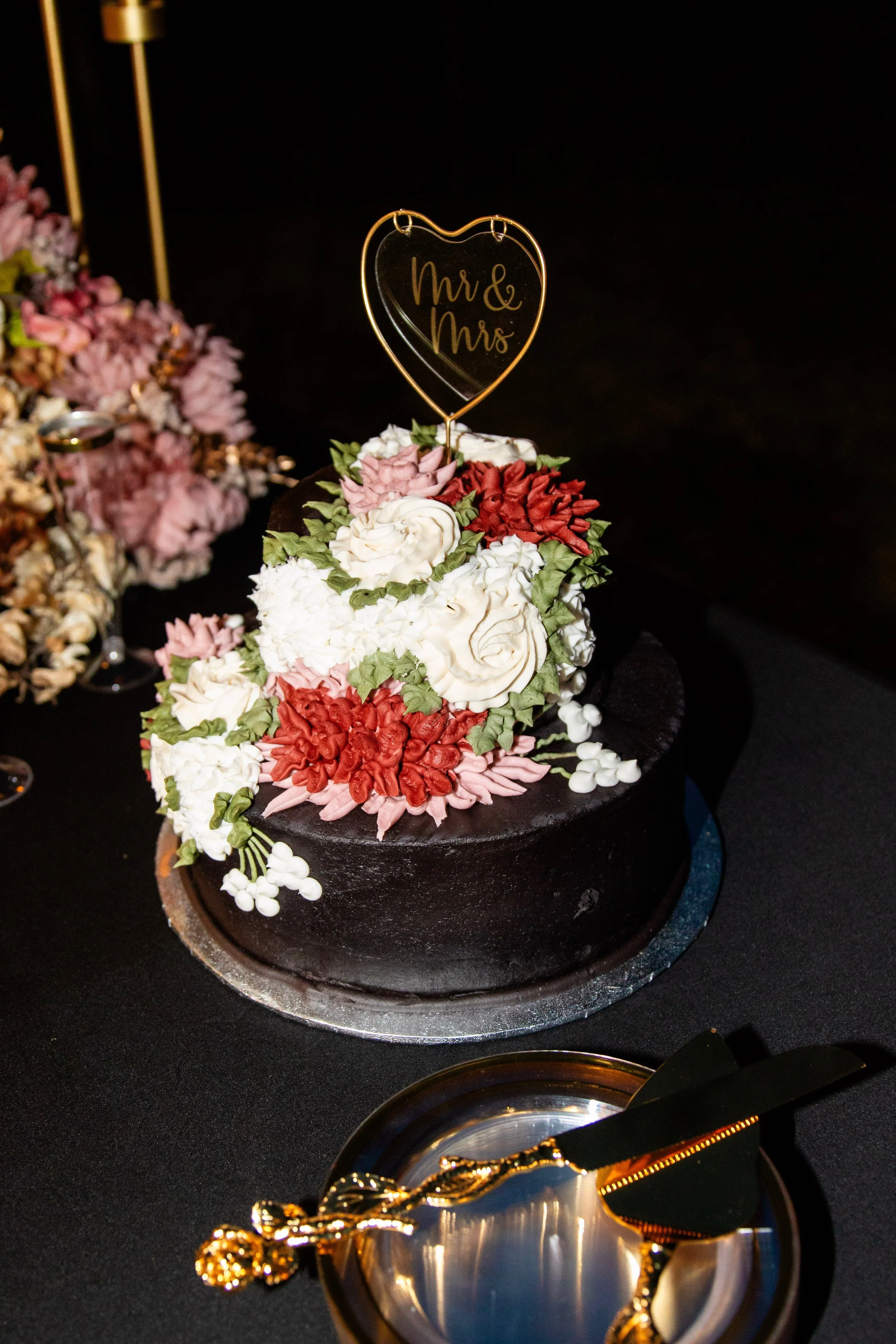 A black wedding cake decorated with white, pink, and red fondant flowers, topped with a heart-shaped sign that reads 'Mr & Mrs'.