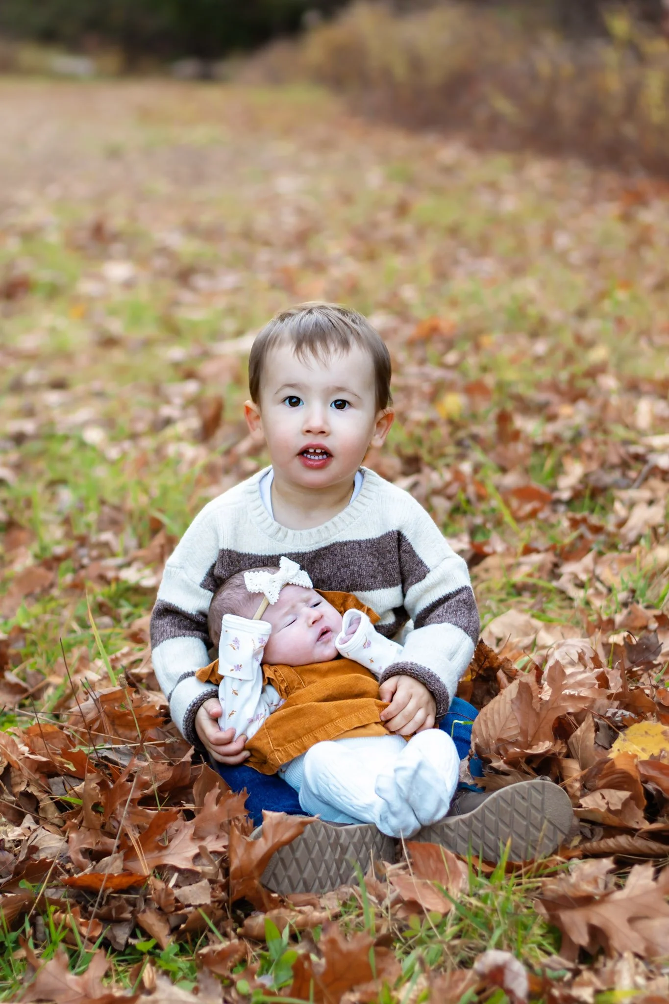 A young boy sitting on fallen autumn leaves in a park, holding a sleeping baby girl in his lap.