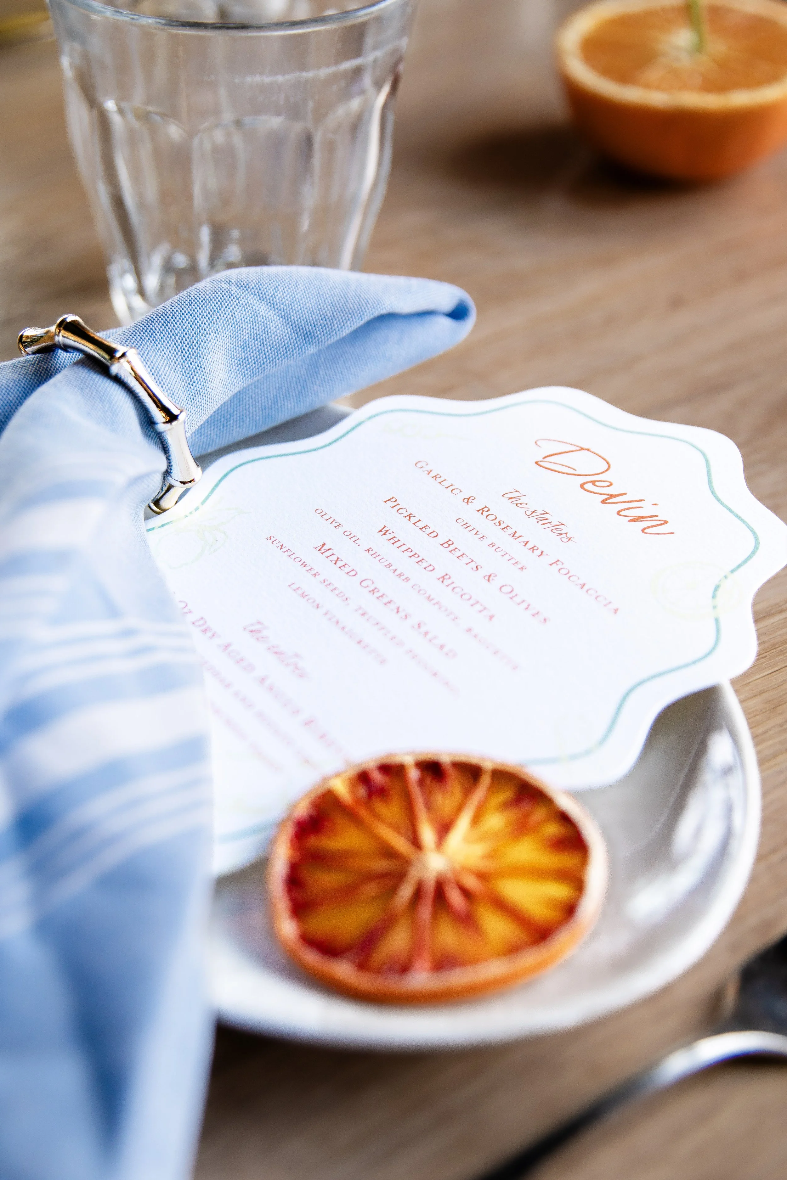 A menu titled "Peonies" with list of ingredients and dishes, a glass of water, half an orange, and dried orange slices on a wooden table.