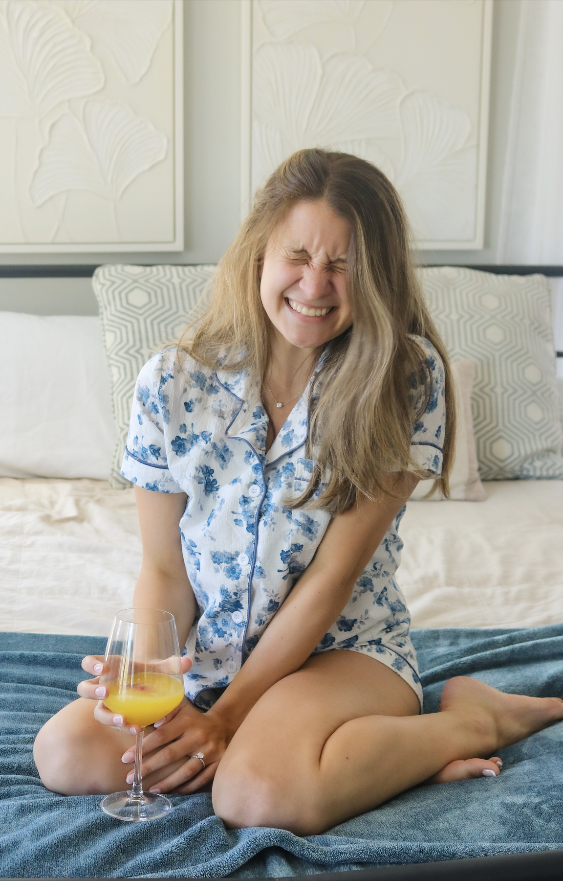 A woman with long hair in pajamas sitting on a bed, grimacing and holding a glass of orange juice.