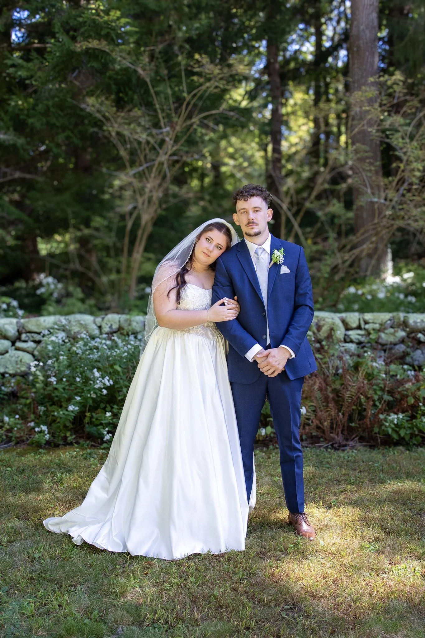 A bride and groom posing outdoors in a wooded area on their wedding day, with the bride wearing a white wedding dress and veil, and the groom dressed in a navy suit with a white shirt and tie.