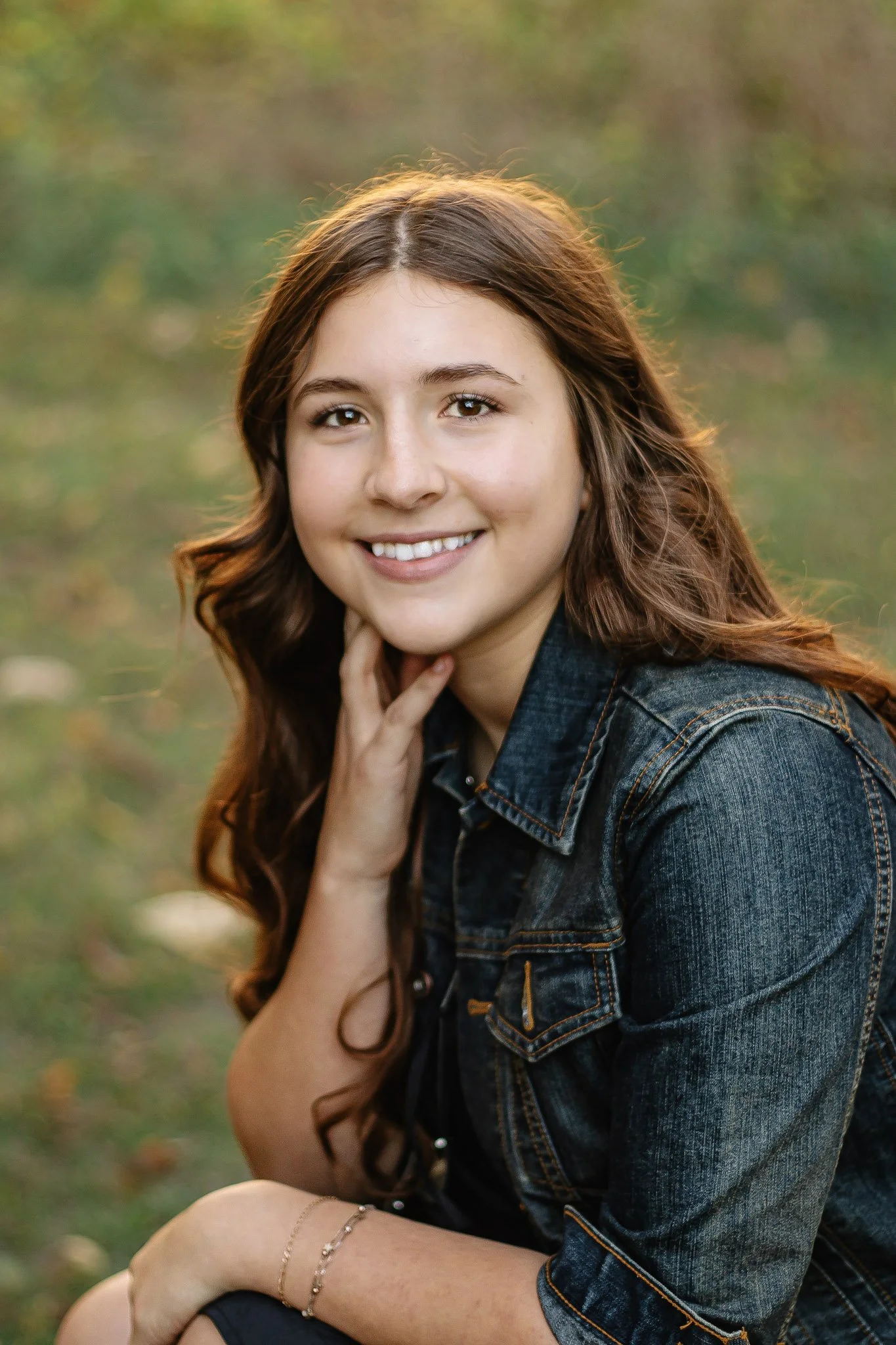 A young woman with long, wavy brown hair smiling outdoors, wearing a denim jacket, with a blurred background of trees and grass.