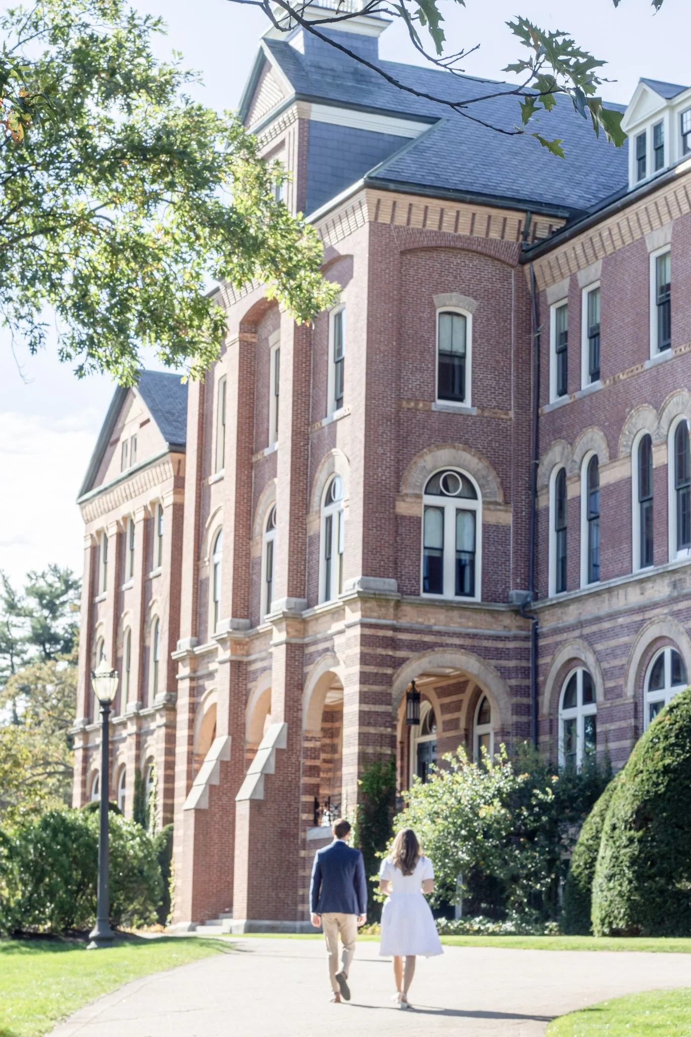 A couple walking towards a large, Victorian-style brick building surrounded by trees and greenery.