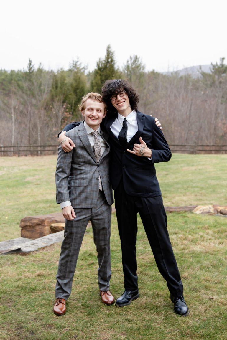 Two men standing outdoors, smiling, with their arms around each other. One is wearing a plaid suit and the other is in a black suit with glasses and curly hair.
