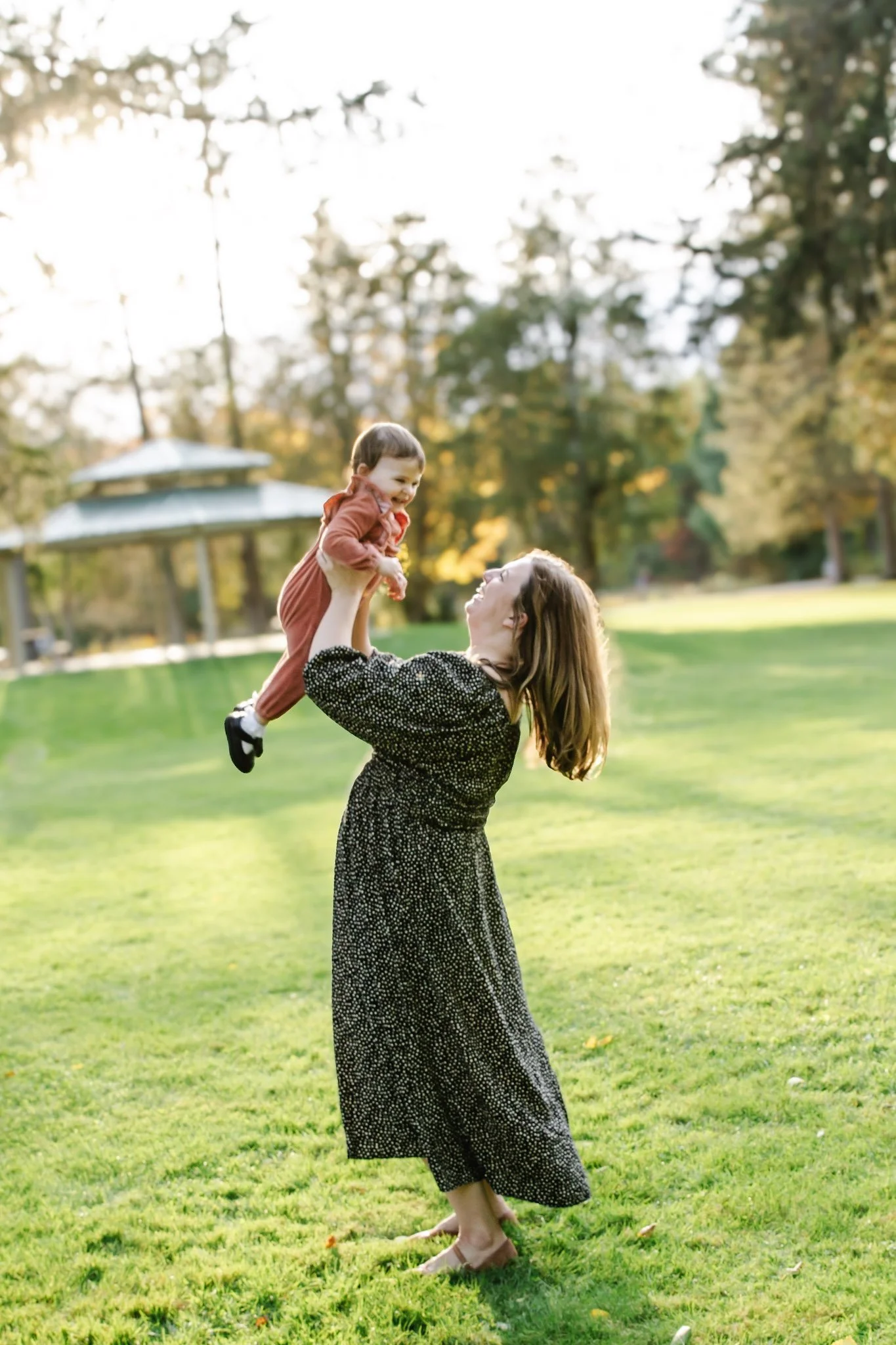 A woman in a black and white dress lifts a young girl in a red dress into the air on a grassy park. The girl is smiling and holding the woman's hands. Green trees and a gazebo are in the background with sunlight filtering through the leaves.