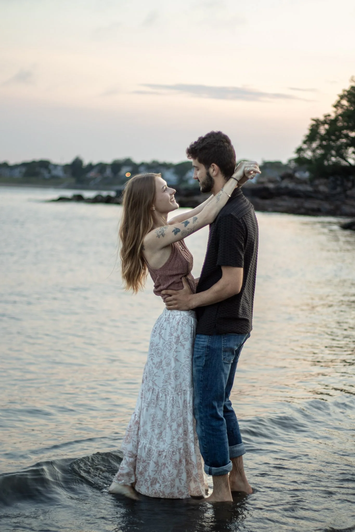 A couple stands in shallow water at a beach during sunset, holding each other and smiling, with trees and houses in the background.