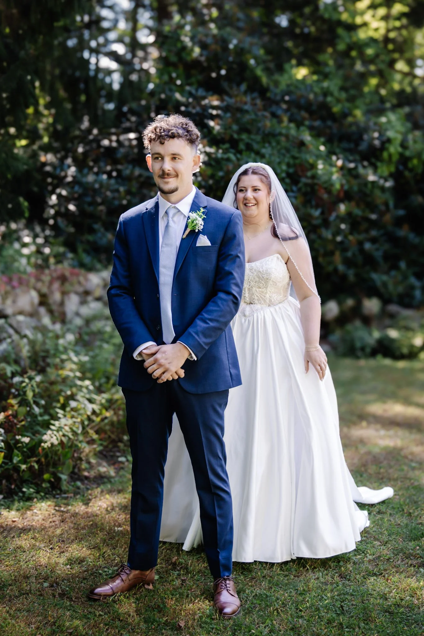 A groom in a navy blue suit and a bride in a white wedding dress standing outdoors with green foliage in the background.