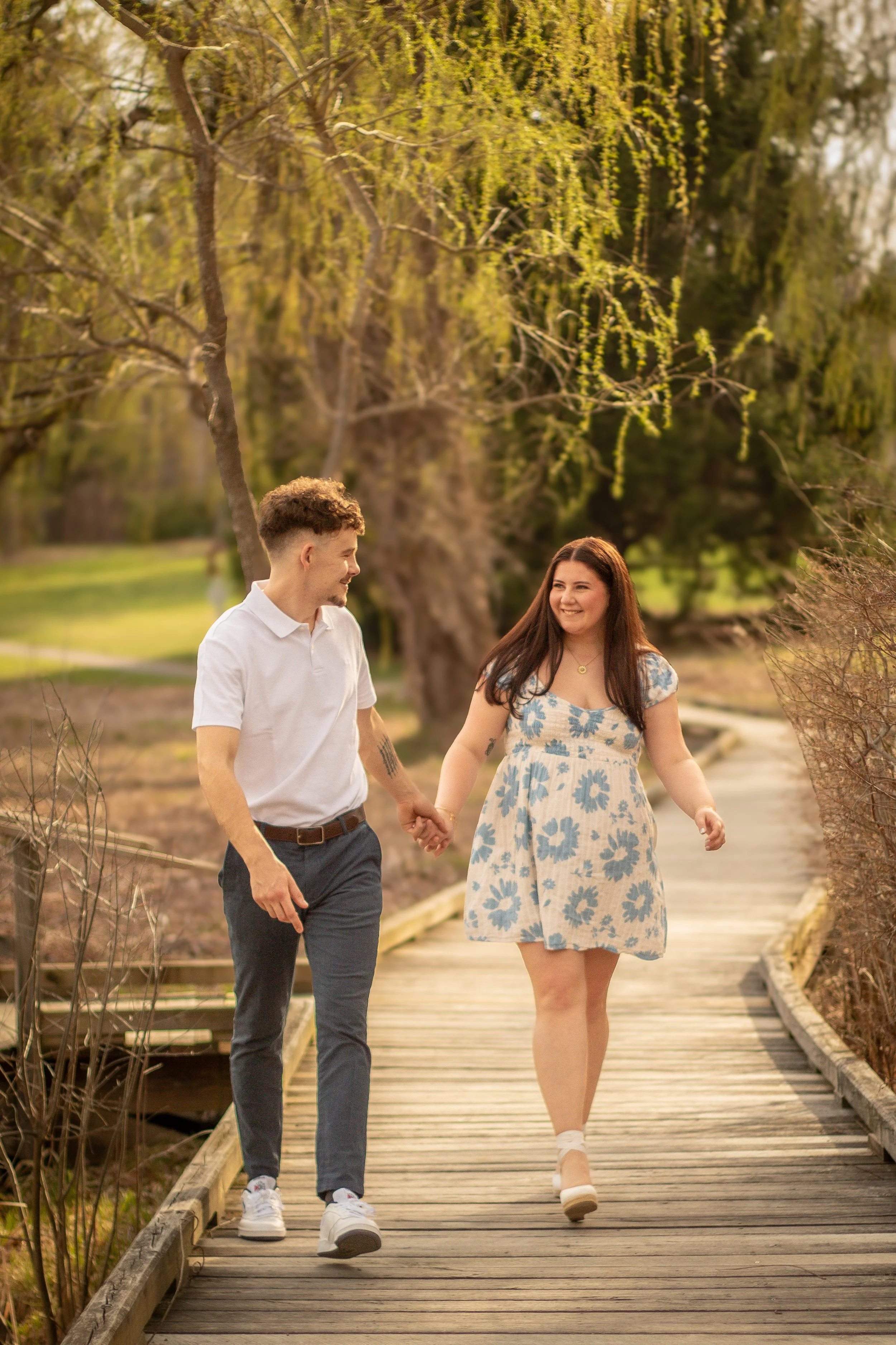 A young couple walking hand-in-hand on a wooden pathway in a park during spring, surrounded by trees and greenery, smiling at each other.