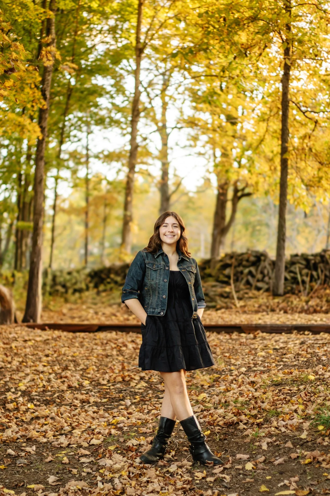 A young woman standing in a park during autumn, surrounded by fallen leaves and yellow-orange trees, smiling and wearing a black dress, denim jacket, and black boots.