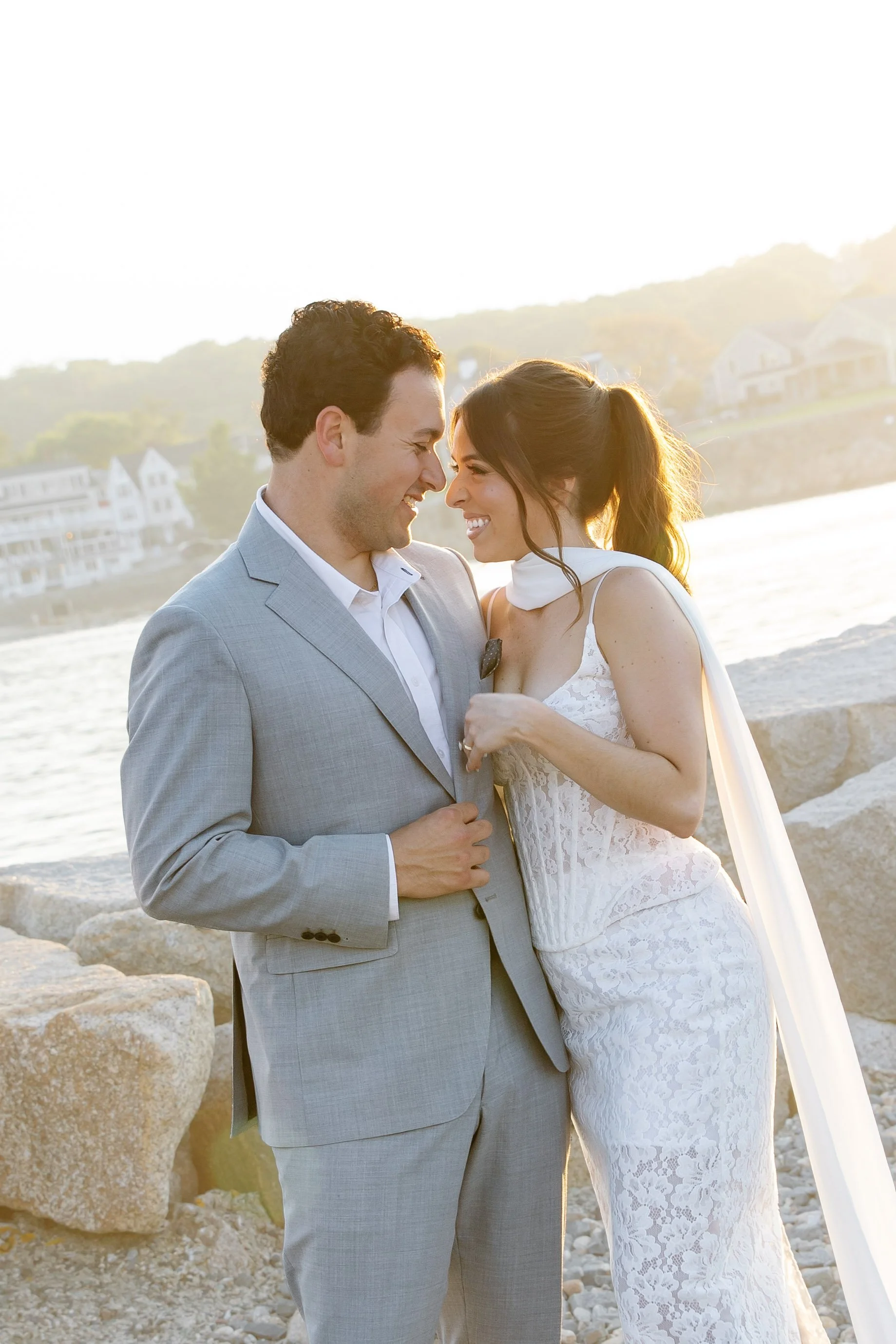 A happy couple, dressed in wedding attire, sharing a moment by the water during sunset, with scenic houses on a hillside in the background.