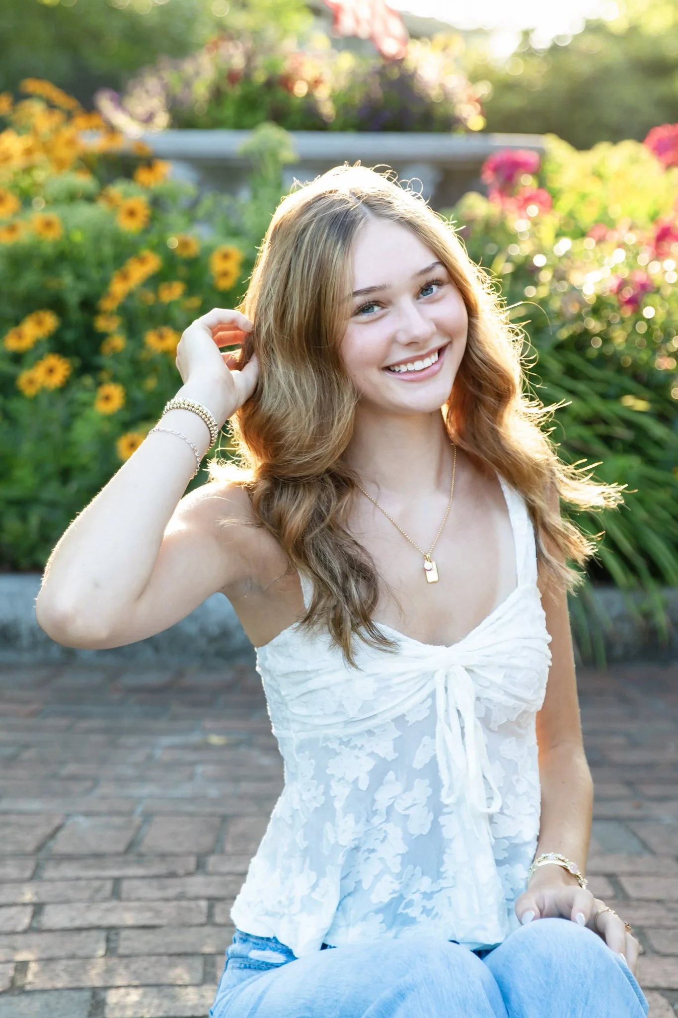 Smiling young woman with wavy light brown hair, wearing a white sleeveless top with lace details, sitting outdoors on brick pavement in a garden with colorful flowers and greenery, backlit by sunlight.