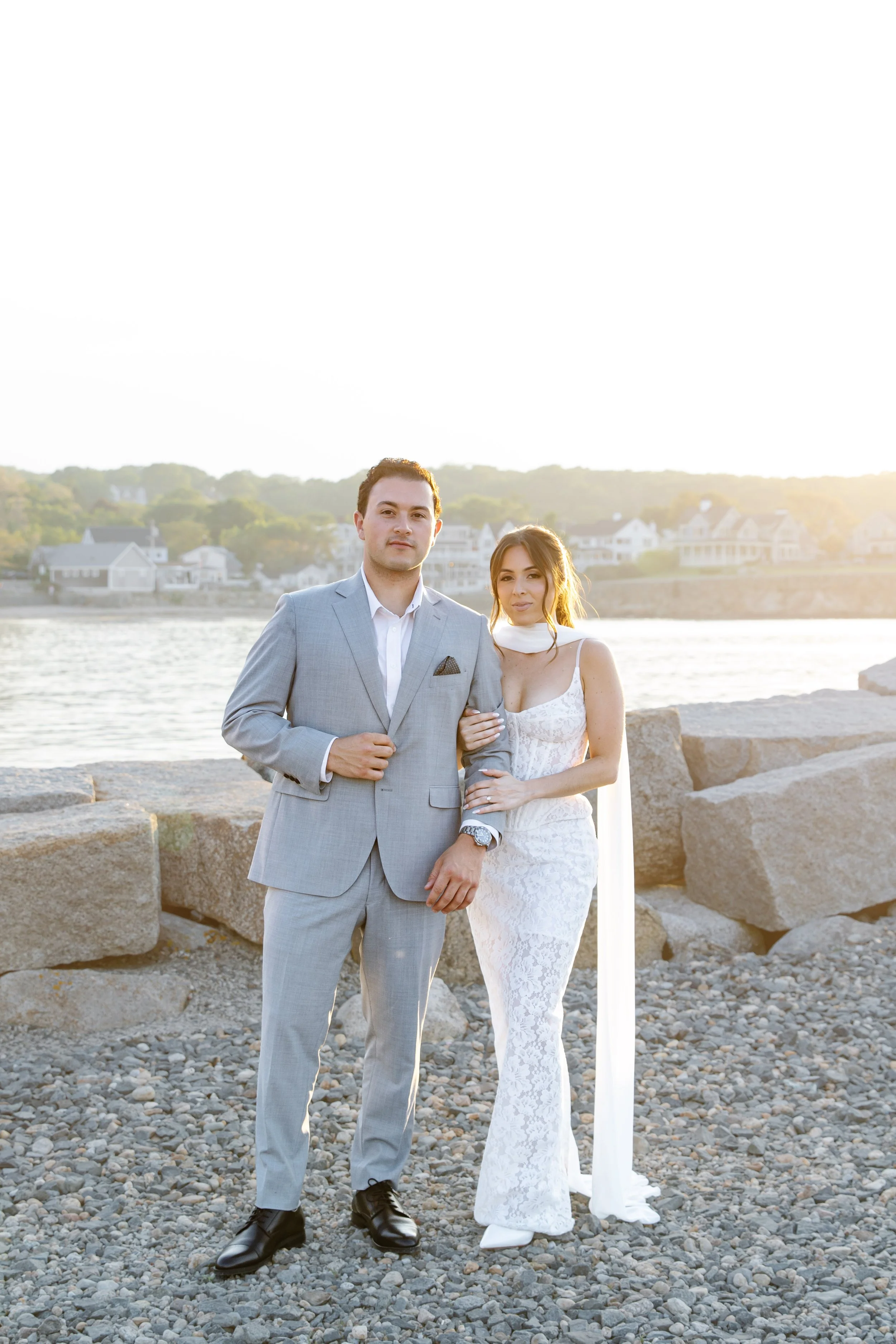 A man and woman standing next to each other outdoors on rocky terrain during sunset. The man wears a light gray suit and the woman wears a white lace dress. The background features water and houses along the shoreline.