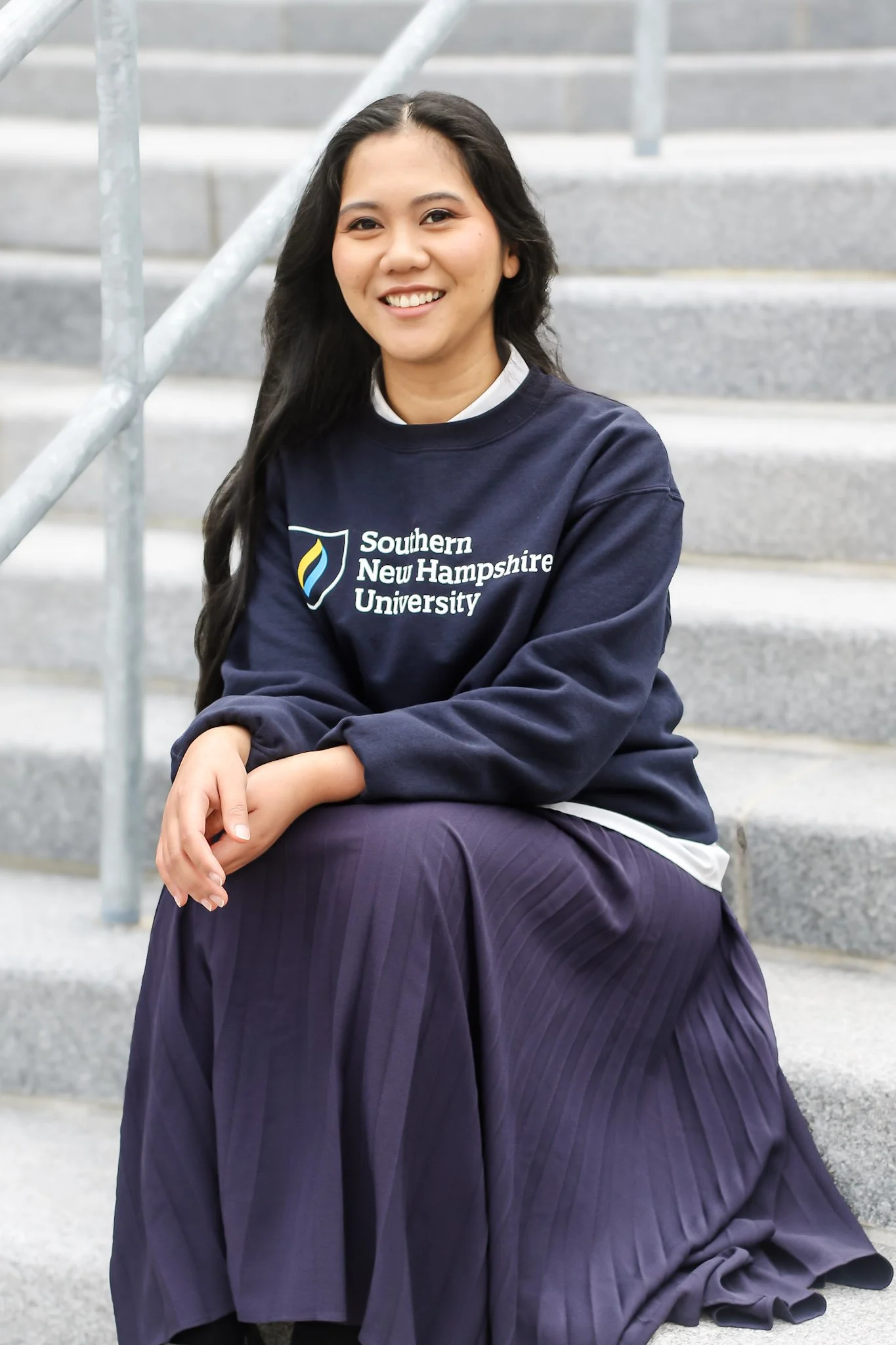 A young woman with long black hair, sitting on steps outdoors, smiling at the camera. She is wearing a navy blue sweatshirt with 'Southern New Hampshire University' written on it and a pleated purple skirt.