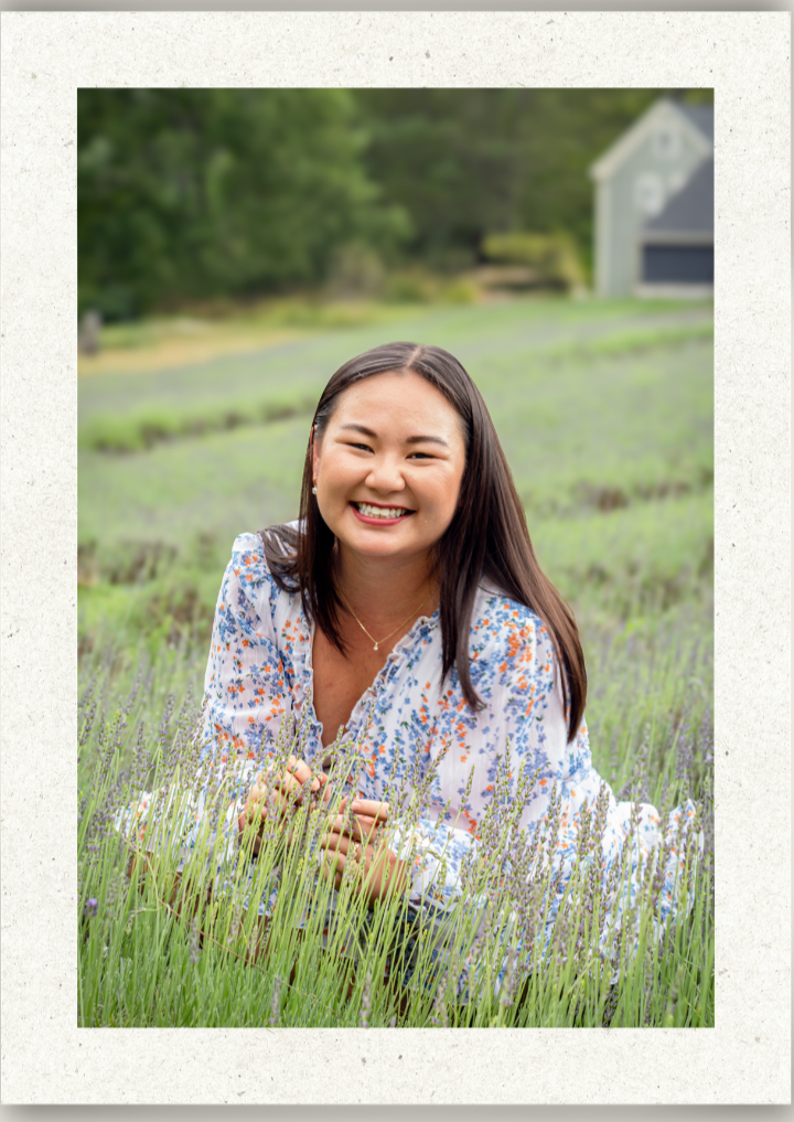 Elena Joy Photography headshots, A woman smiling and lying in a field of lavender, with a house and trees in the background.