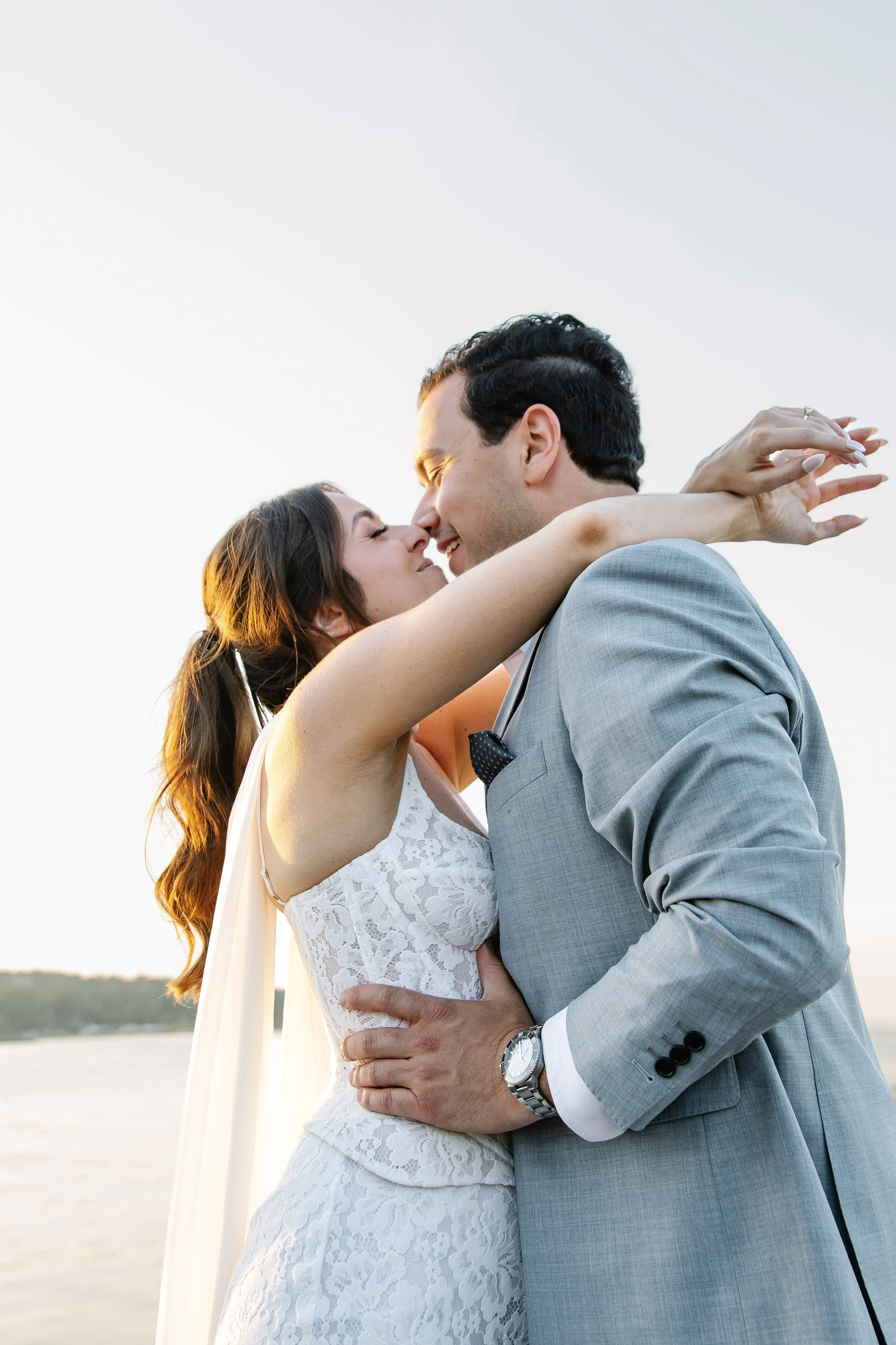 A newlywed couple embraces by the water during sunset, with the bride in a white lace dress and the groom in a light gray suit, both smiling and about to kiss.
