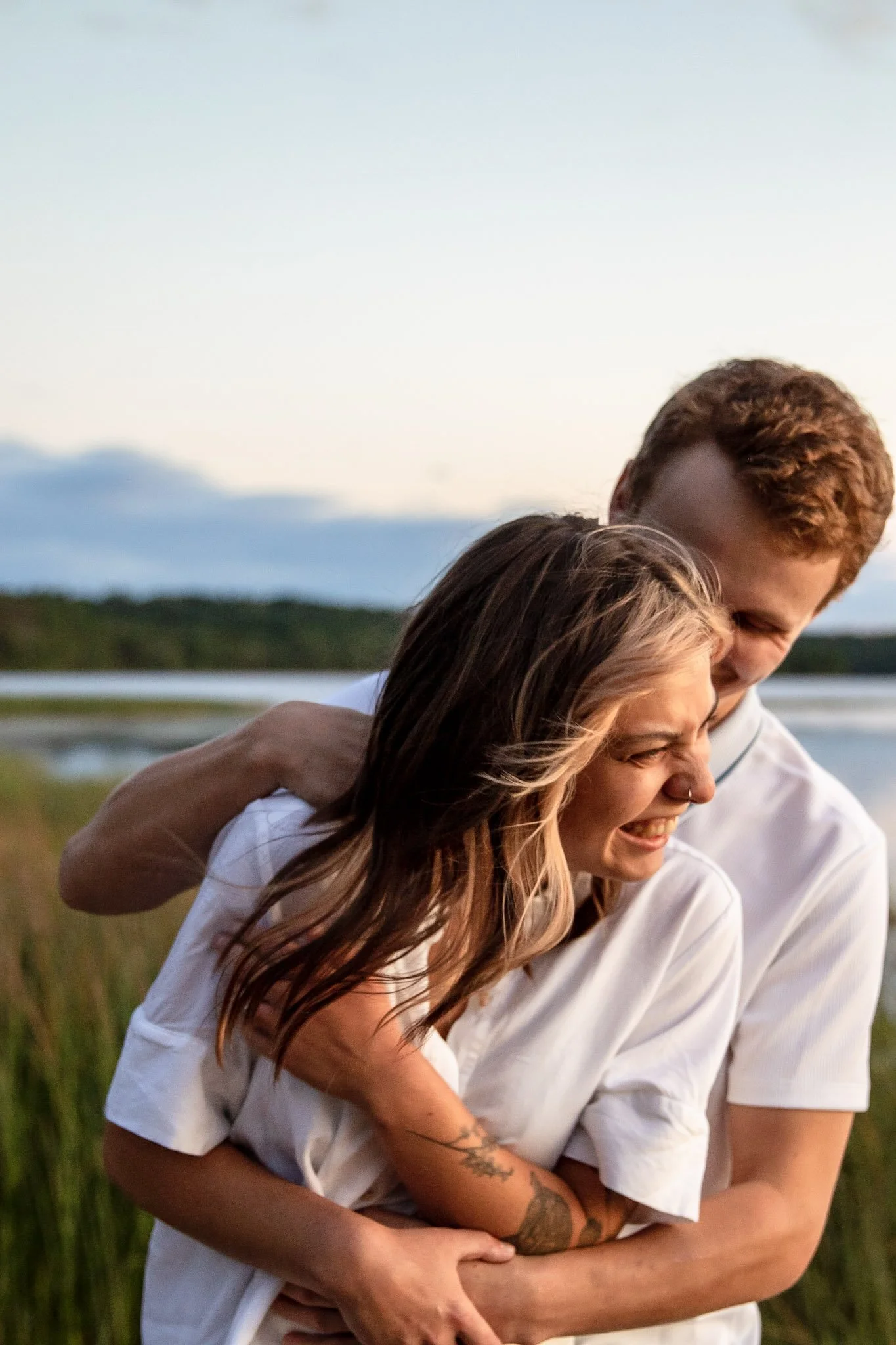 A young couple sharing a joyful embrace outdoors by a water body during sunset.