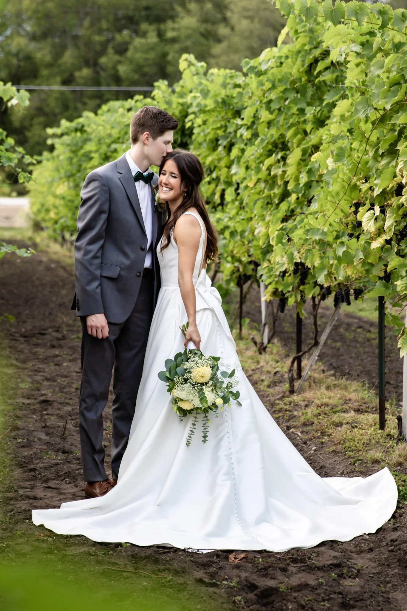 A newlywed couple stands close together in a vineyard, with the groom gently kissing the bride's forehead. The bride is in a white wedding gown holding a bouquet of white and green flowers, smiling happily, while the groom is in a gray suit with a bl