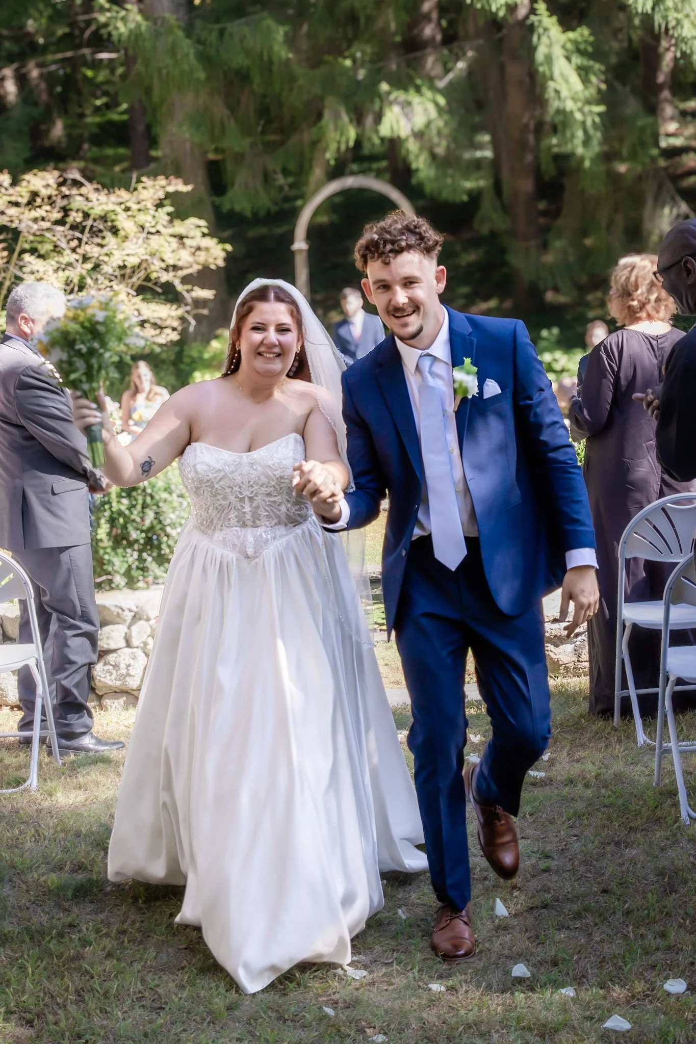 A newlywed couple smiling and holding hands, walking outdoors at their wedding reception in a forested area, with guests in the background.