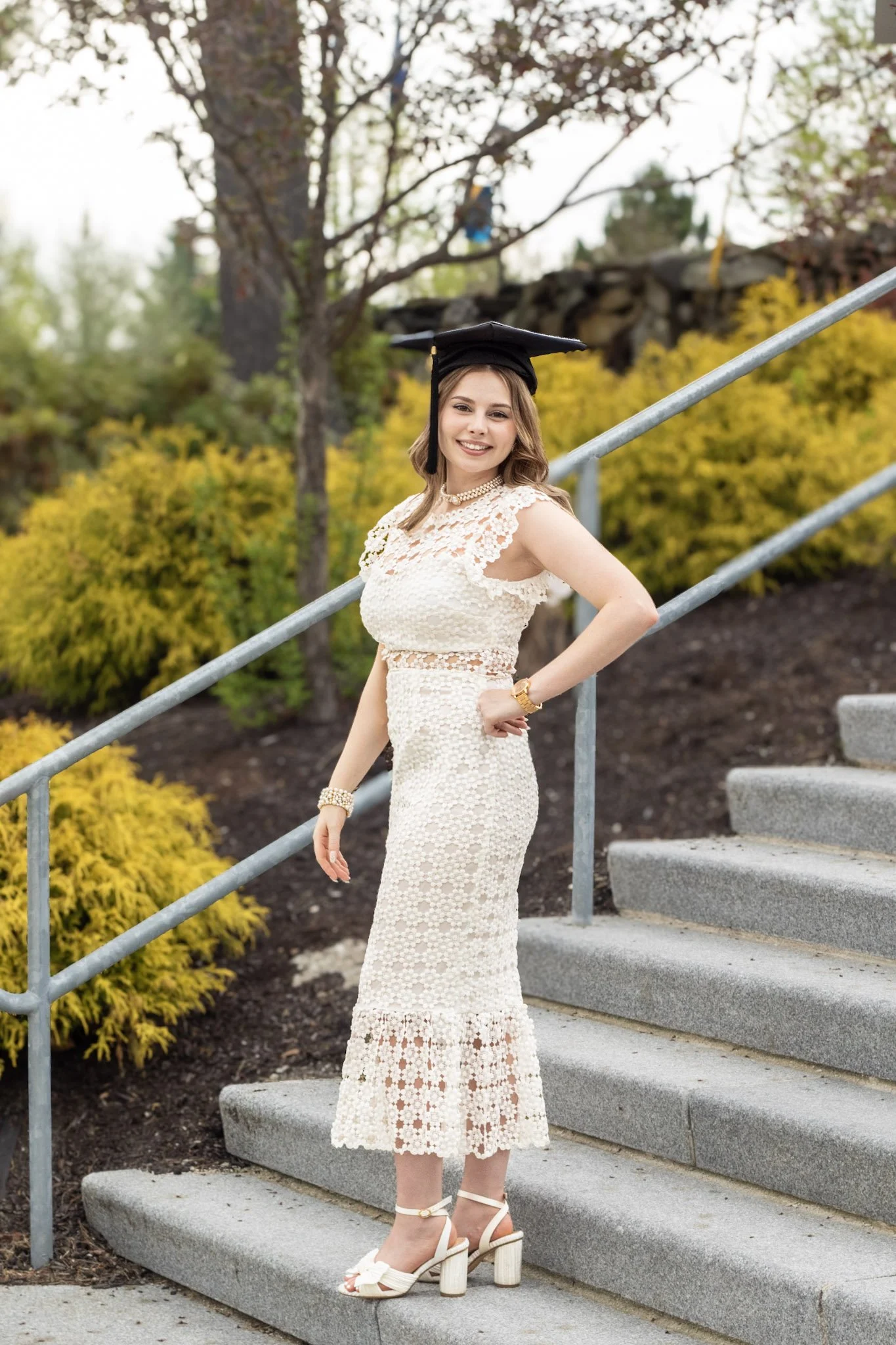 A young woman in a white lace dress, wearing a black graduation cap, is standing on outdoor steps and smiling at the camera.