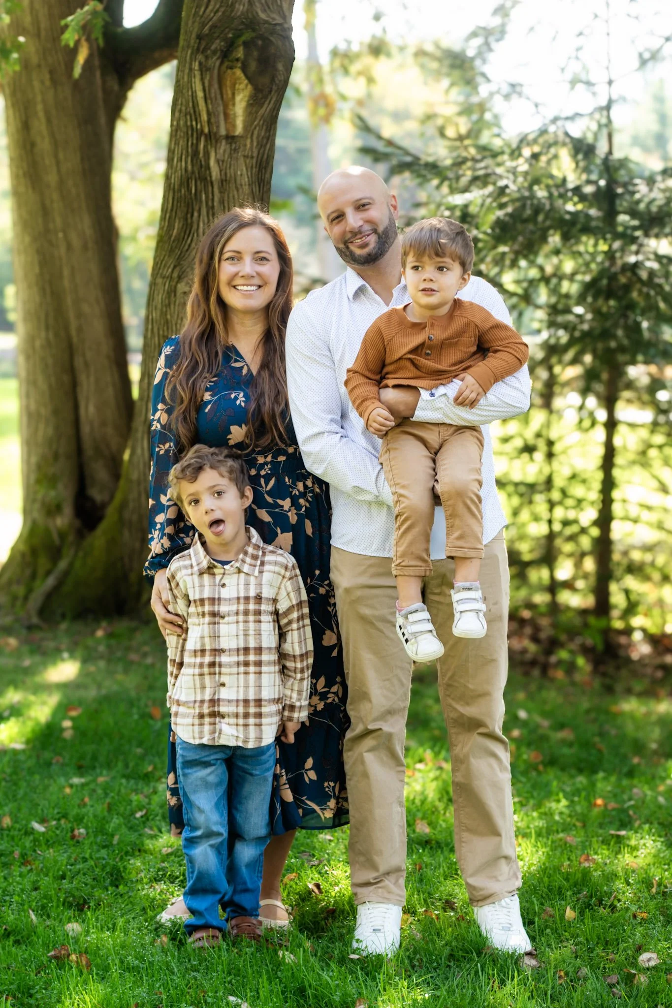 Benson Park, NH, Family of five standing outdoors in a park, smiling and posing for a photo. They are surrounded by green grass and trees, with sunlight filtering through the leaves.