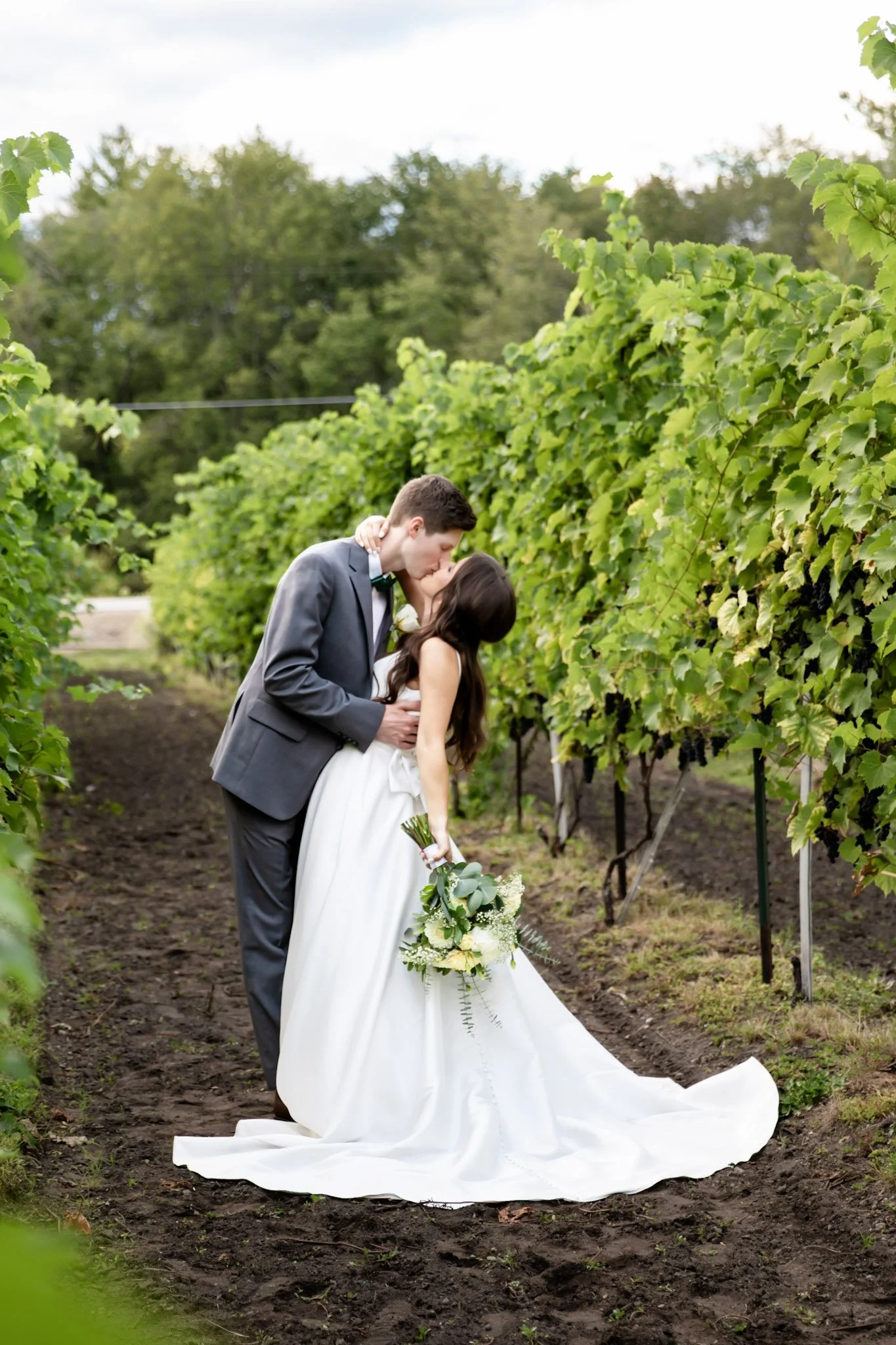 A bride and groom kissing in a vineyard, with the bride holding a bouquet of flowers.