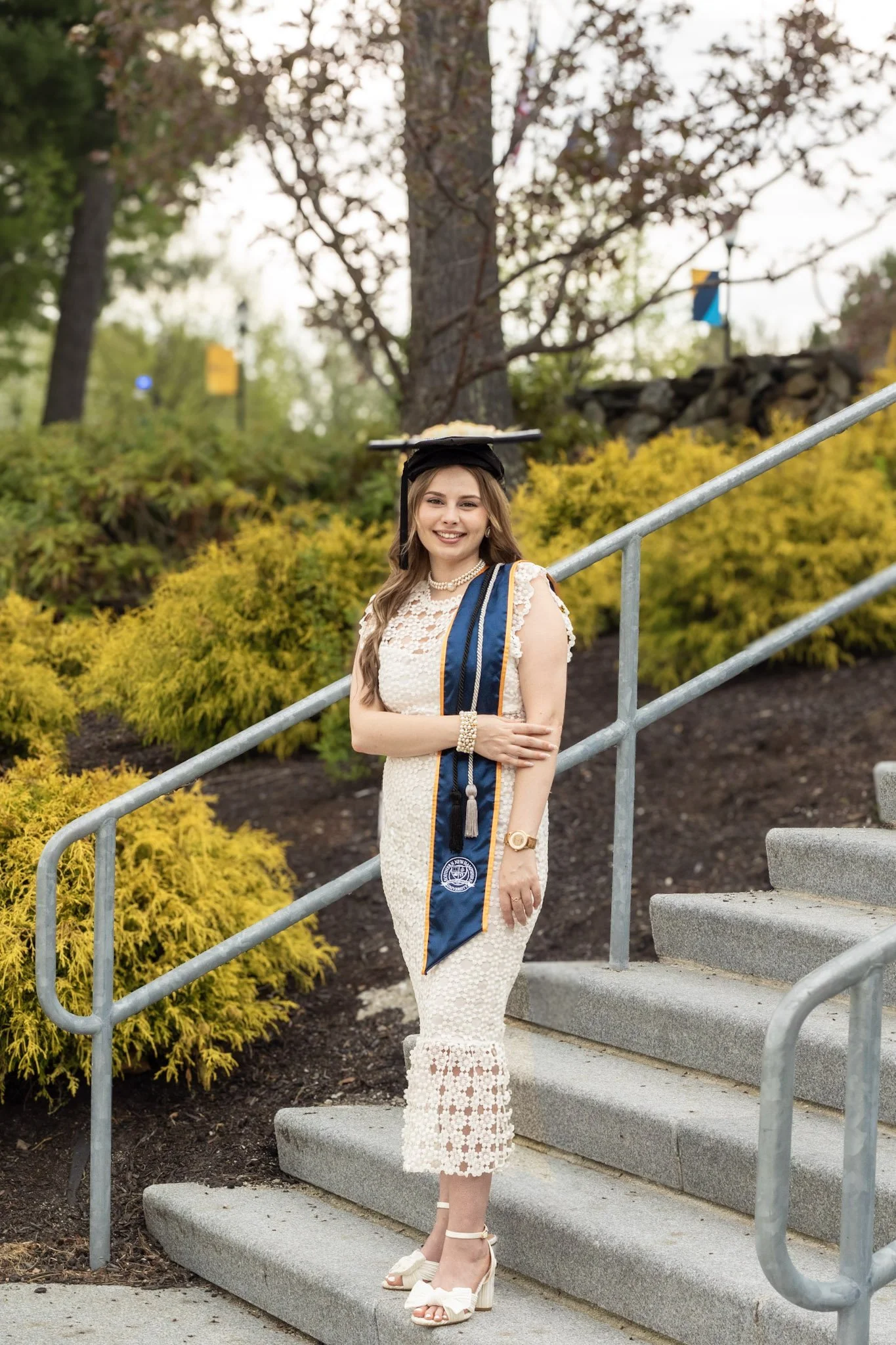 A young woman in a white lace dress, wearing a graduation cap and gown, stands on outdoor stairs smiling with her arms crossed, surrounded by yellow bushes and trees.