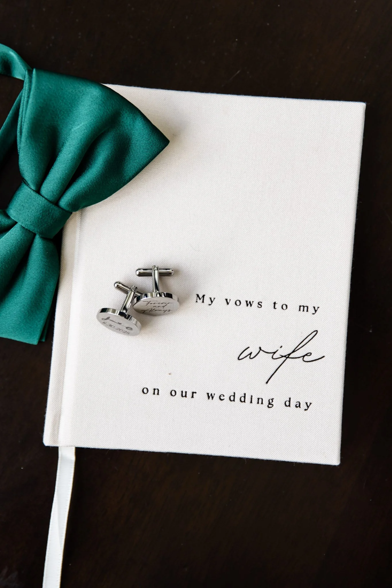 Wedding vows notebook with a teal bow tie and silver cufflinks on top, placed on a dark wooden surface.