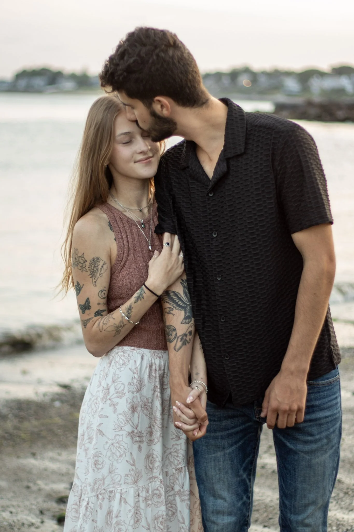 A young couple with tattoos holding hands and sharing an intimate moment on the beach, with water and distant shoreline in the background.