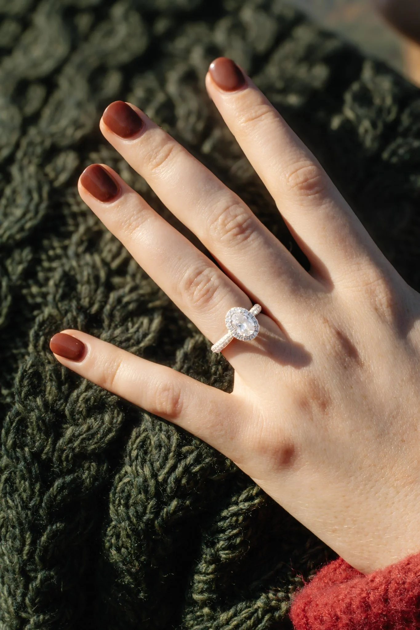 A person with painted nails displays a ring with a large oval gemstone surrounded by smaller stones on their ring finger. They are wearing a red sleeve and are resting their hand on a textured green woven surface.