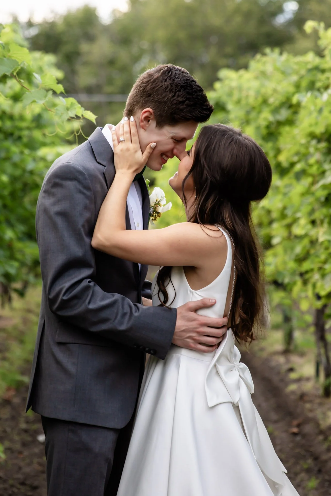 A couple in wedding attire sharing a romantic moment outdoors, with their faces close, surrounded by lush greenery.