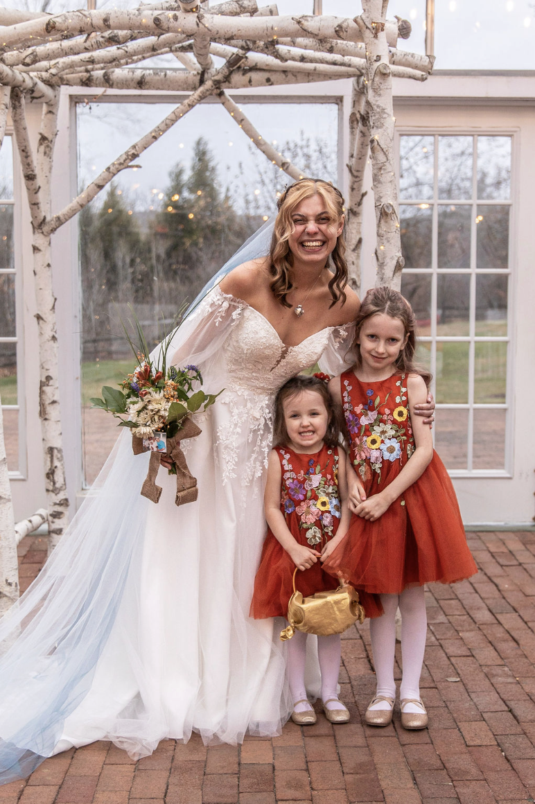 A bride in a white wedding gown smiling with two young girls wearing red dresses with floral embroidery, standing under a rustic arbor made of birch branches outdoors.