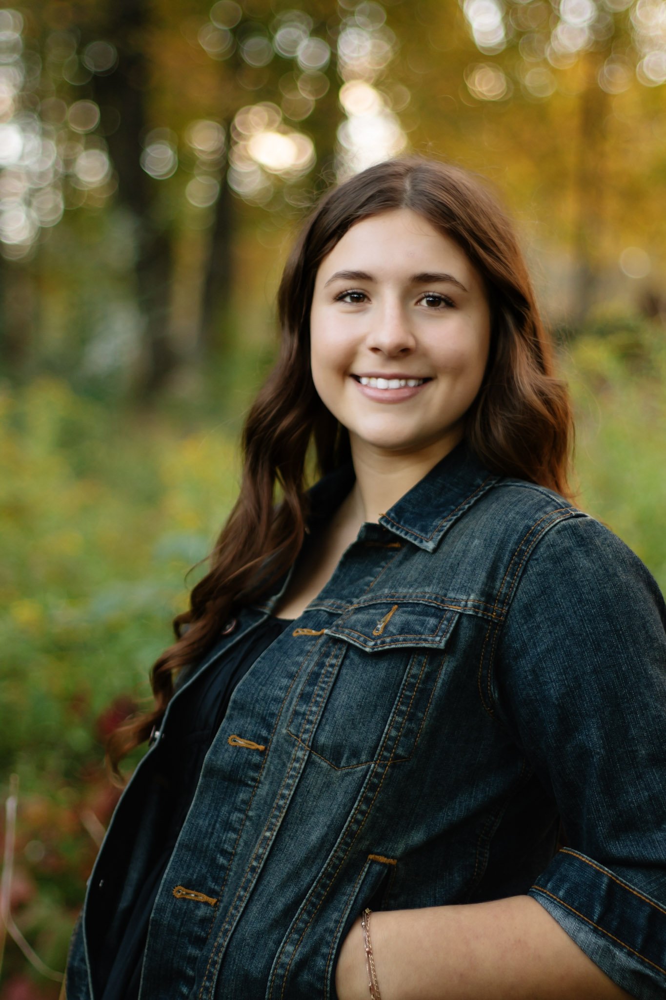 Smiling woman with long brown hair in a denim jacket standing outdoors in a forest with colorful autumn trees in the background.