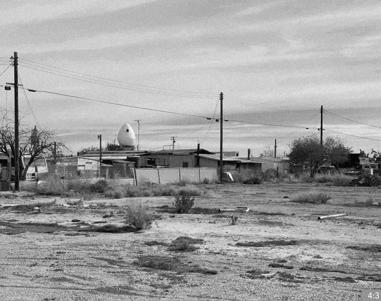 62. Salton Sea 1. SHOT LONG MEDIUM CLOSE: father stands or crouches in the middle of the street alone amongst the shacks and trailers.
6. LONG SHOT: father and daughter stand in the middle of the street looking at one another.