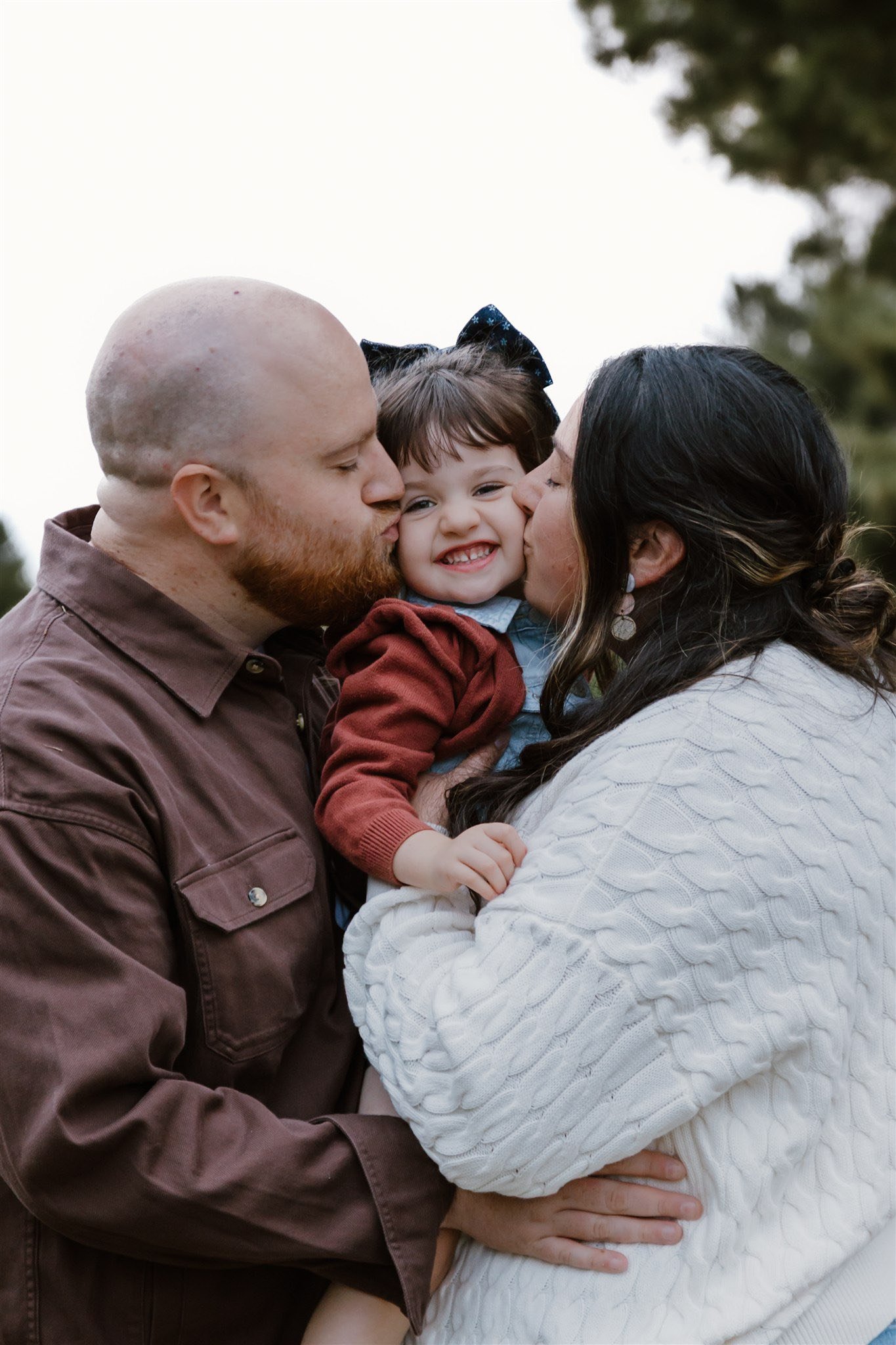 A family of three, a man, a woman, and a young girl, sharing a joyful moment outdoors. The man and woman are kissing the girl on her cheeks, and she is smiling happily.