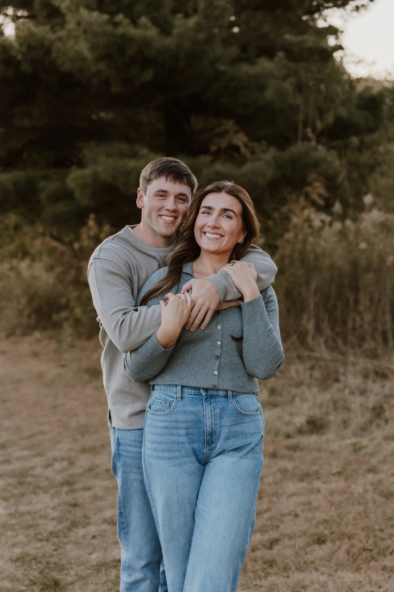 A young couple smiling and embracing outdoors in a natural setting with trees and grass.