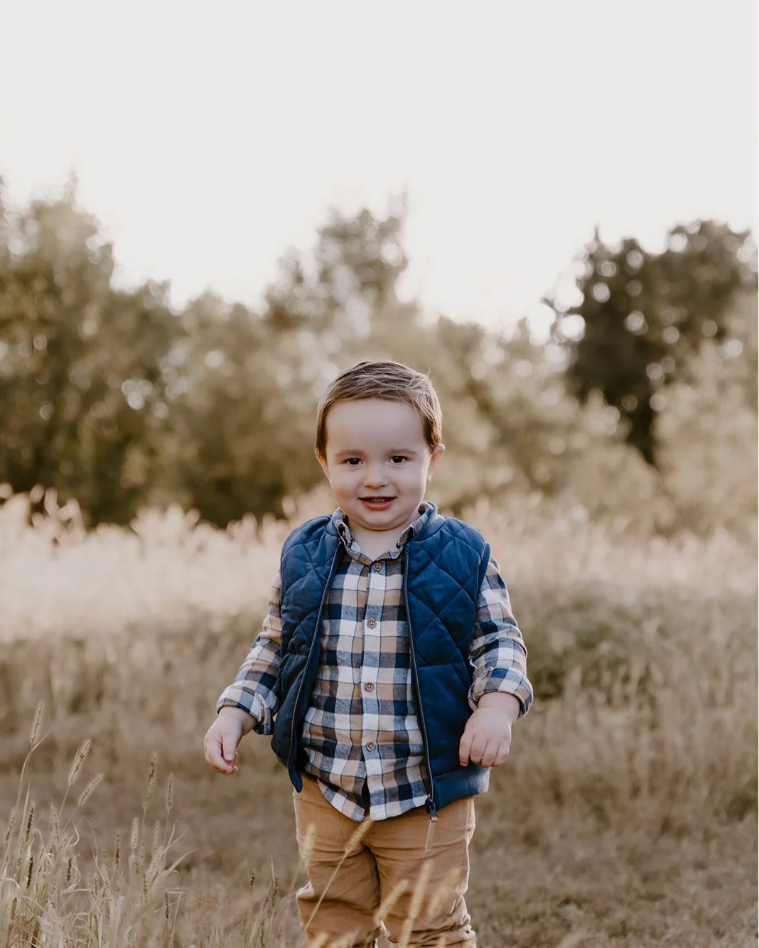 A young boy with brown hair, wearing a blue quilted vest over a plaid shirt and tan pants, walking through a grassy field with trees in the background during daytime.