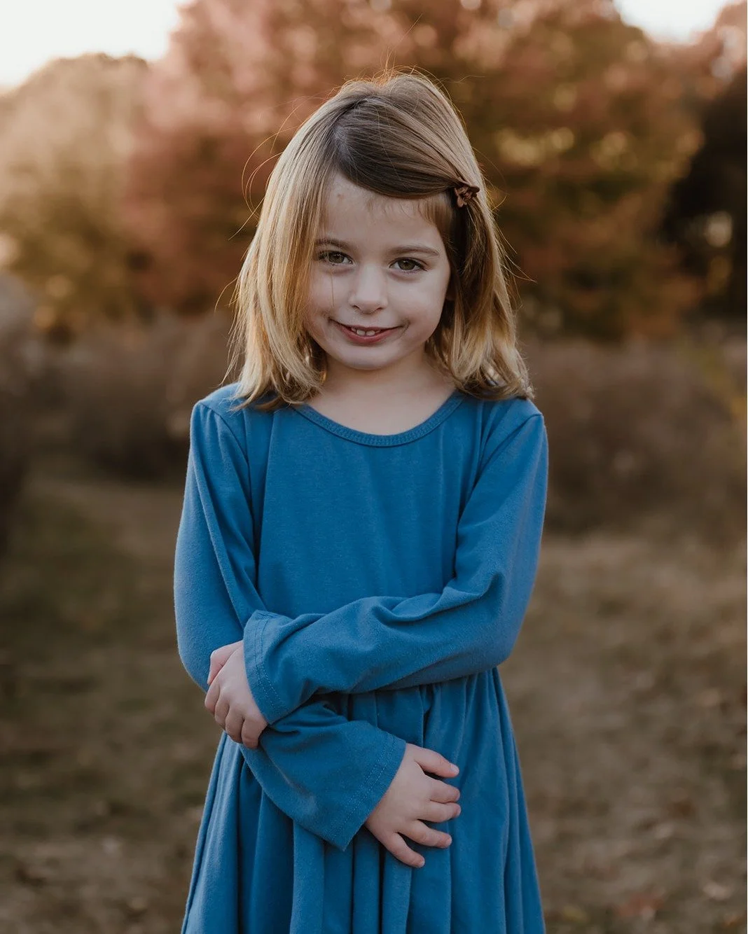 A young girl with blonde hair wearing a blue long-sleeve dress, smiling with her arms crossed, standing outdoors with autumn-colored trees in the background.