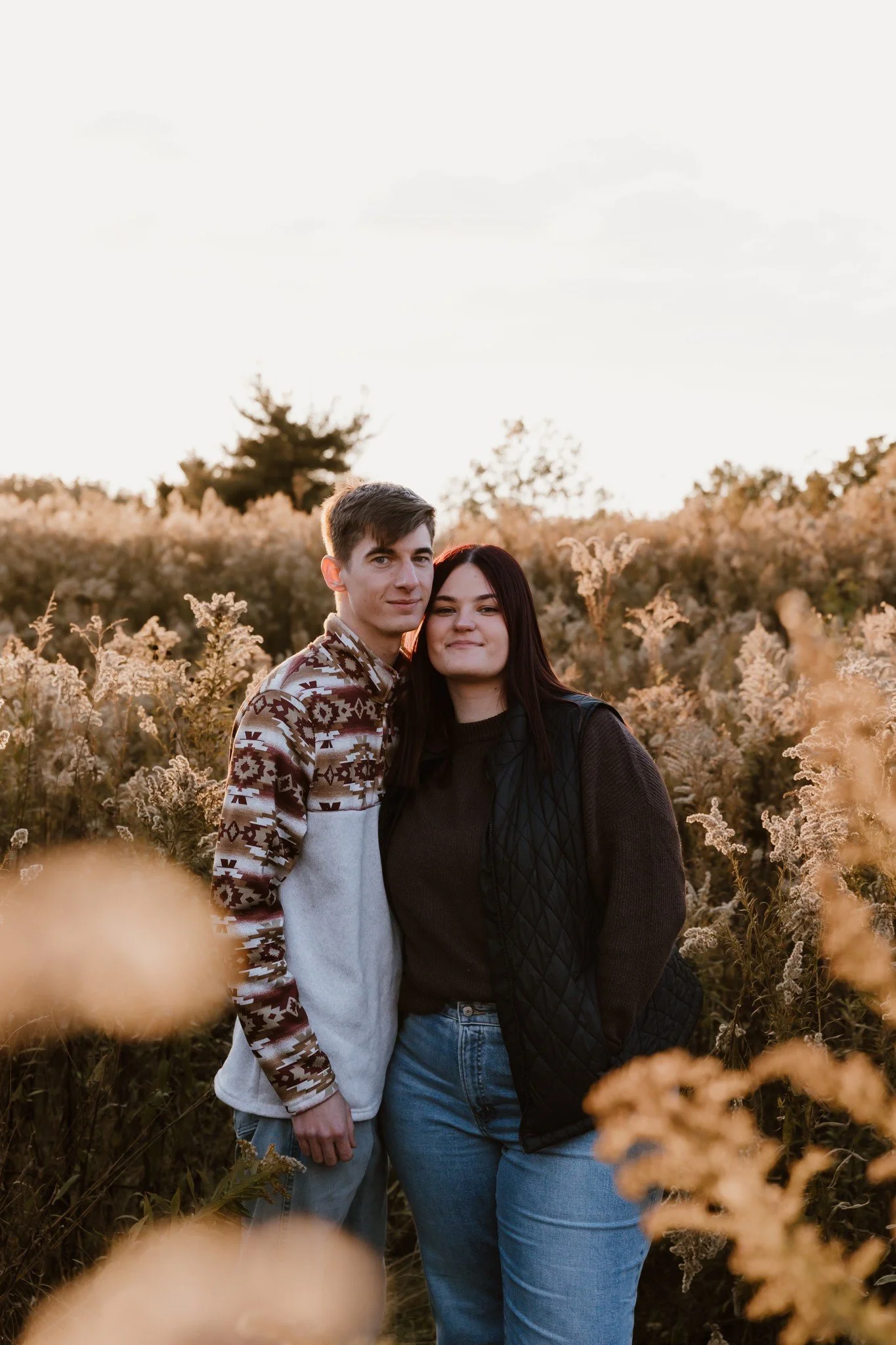 A young couple standing close together in a field of tall, beige and white wildflowers at sunset, with a soft, golden light illuminating their faces.
