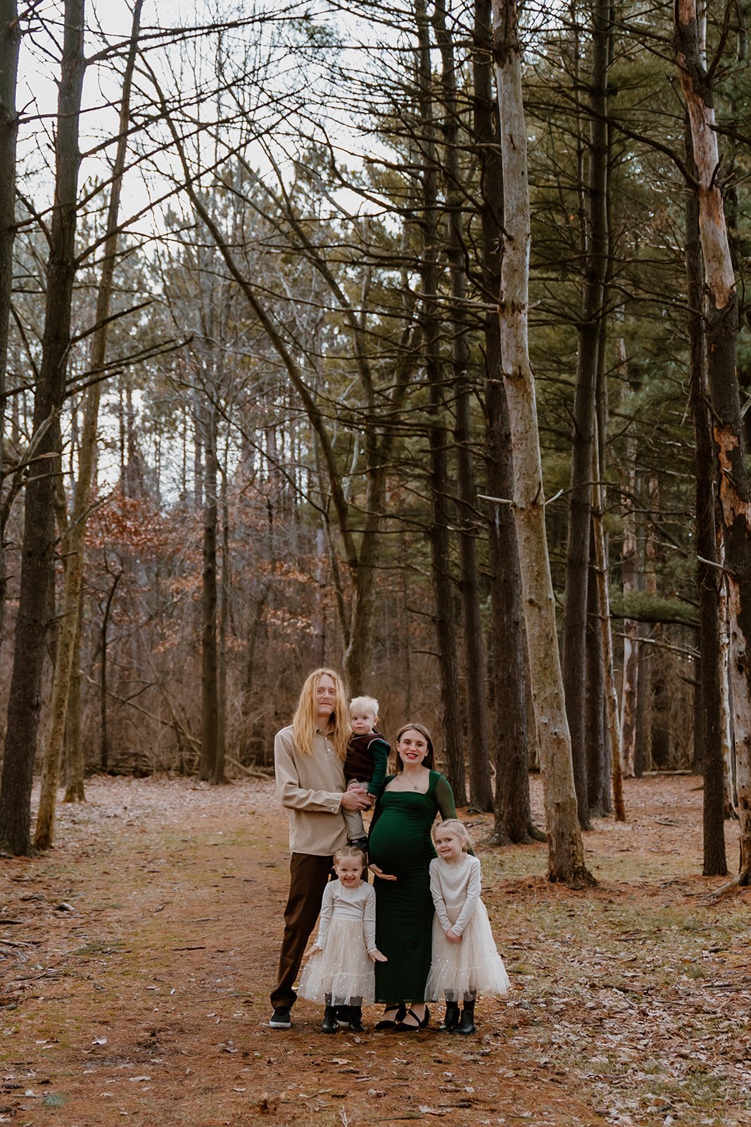 A family of five standing in a wooded forest, with a pregnant woman in a green dress, a man, and three young girls dressed in white dresses, during daytime.