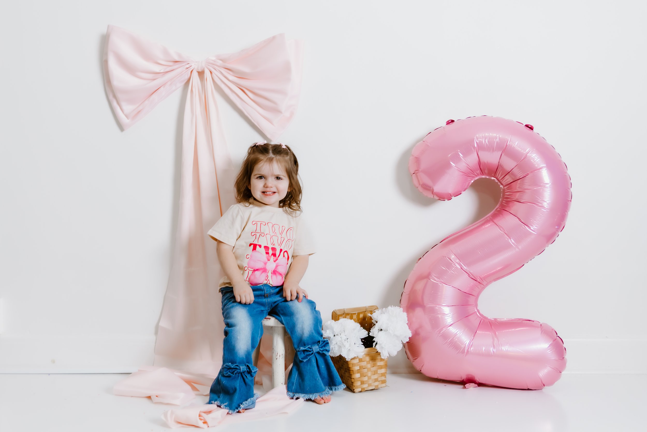 Young girl sitting on a white chair in front of a white wall with pink decorations, including a large pink bow and a pink number two balloon