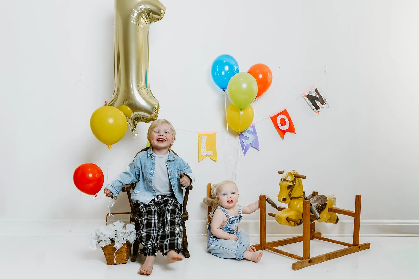 Two young children celebrating a first birthday with balloons, a large gold number one balloon, a banner that spells 'LISO', and a yellow rocking horse in front of a plain white wall.