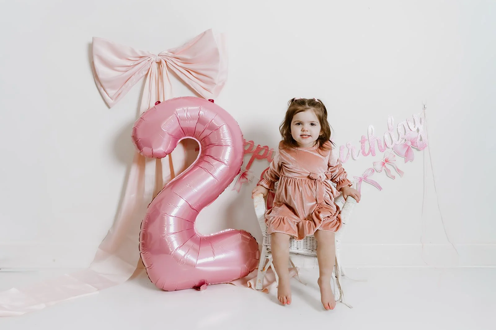 Young girl at a birthday celebration sitting in a white woven chair with pink dress, next to a large pink number two balloon and pink birthday decorations including a bow and "happy birthday" banner.
