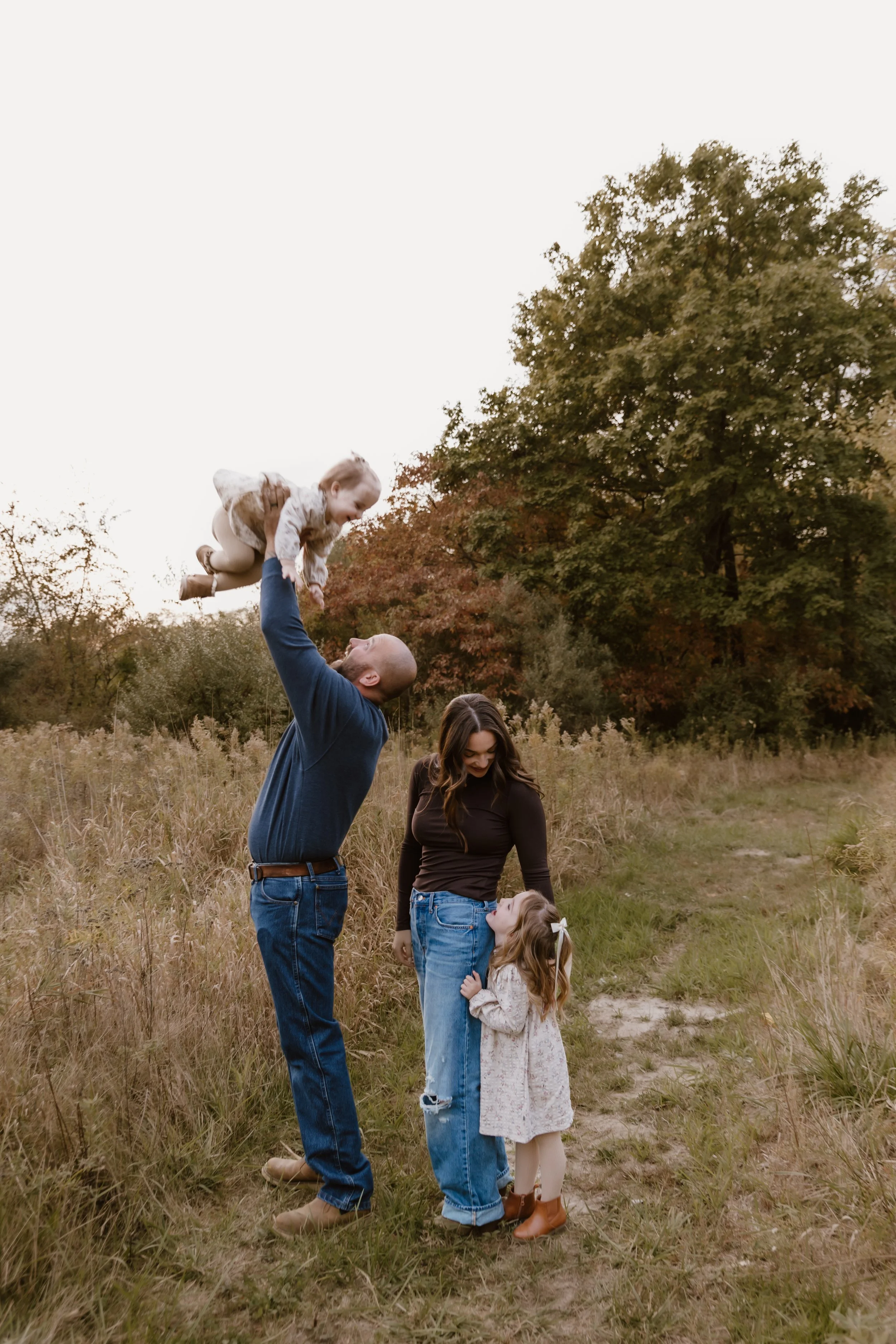 A family of four enjoying time outdoors, with a man lifting a young girl into the air, a woman and a little girl standing nearby in a grassy field during autumn.